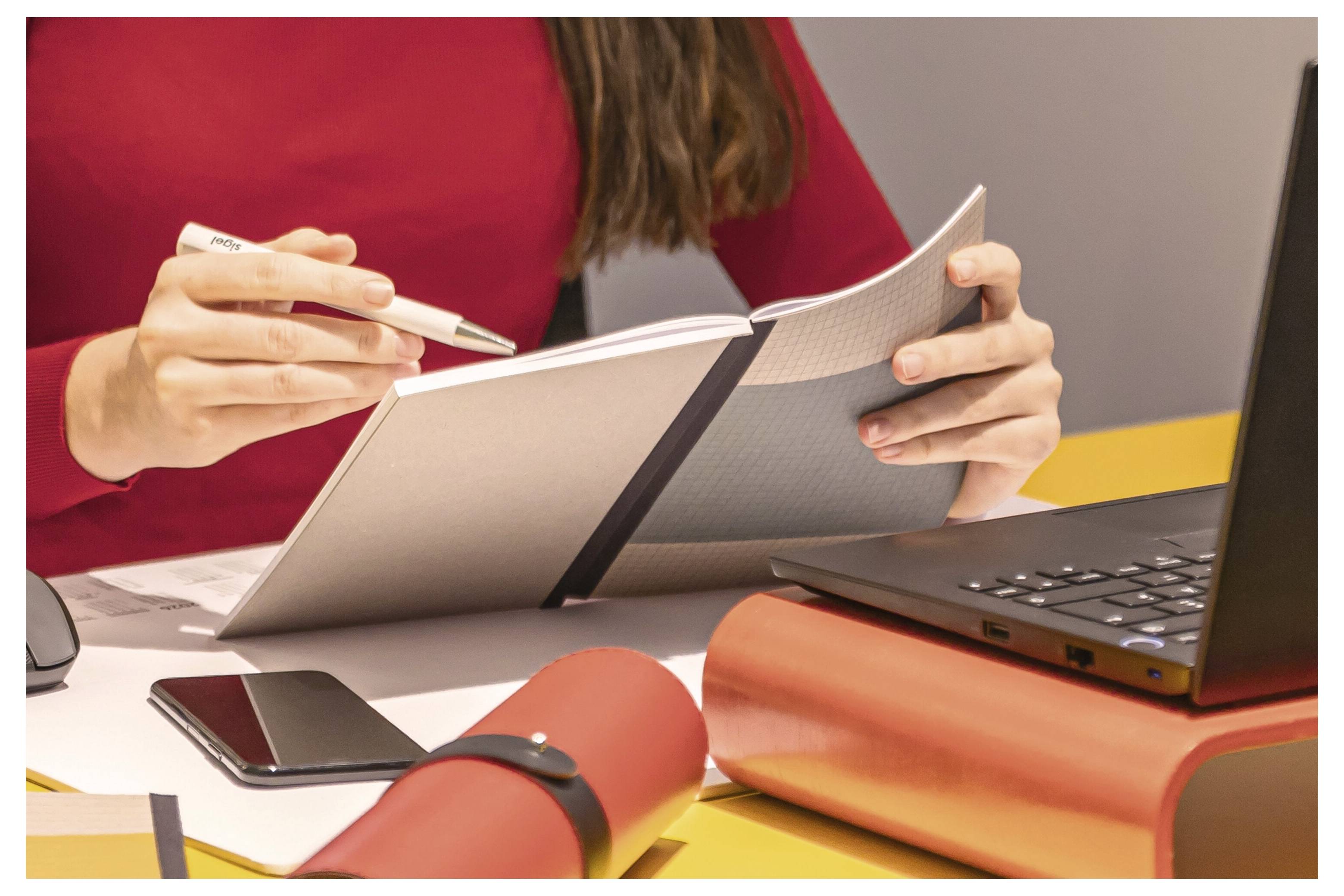 Person in einem roten Oberteil schreibt in einem Notizbuch neben einem Laptop, Smartphone und aufgerollten Dokumenten auf einem Schreibtisch.