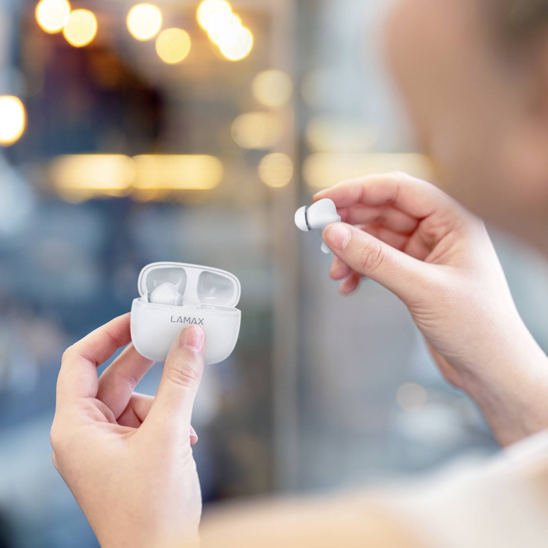 Person hält kabellose Ohrhörer in der Hand, während sie das Ladeetui öffnet. Hintergrund unscharf mit Lichtern.