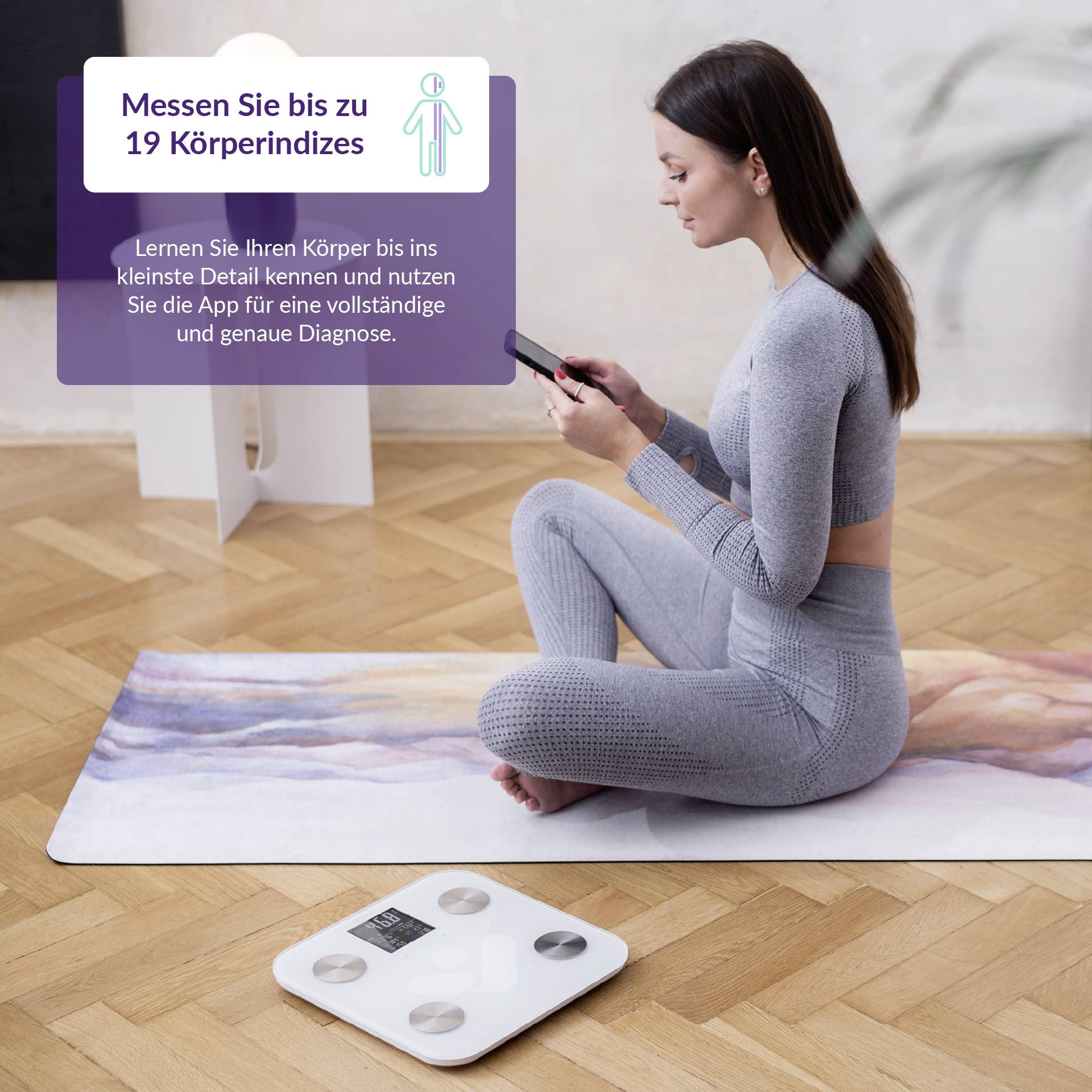 Eine Frau in Sportkleidung sitzt auf einer Yogamatte, hält ein Smartphone. Daneben eine Waage auf dem Boden.