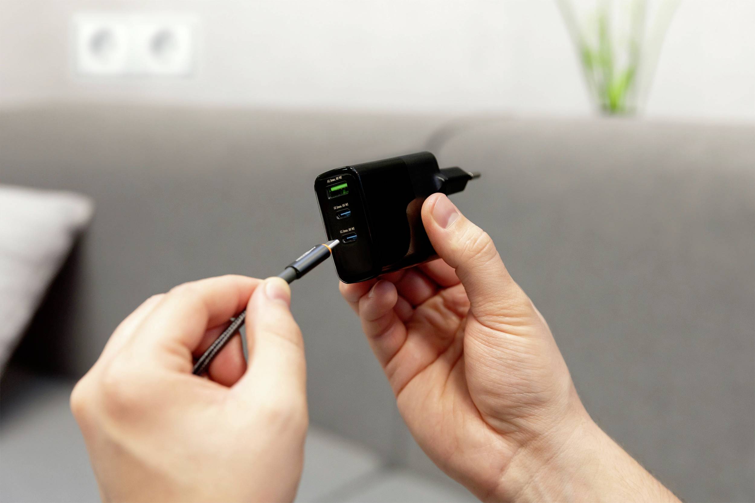 Eine Person hält ein Ladegerät mit mehreren USB-Anschlüssen in der Hand und steckt ein Kabel ein. Im Hintergrund ist ein Sofa zu sehen.