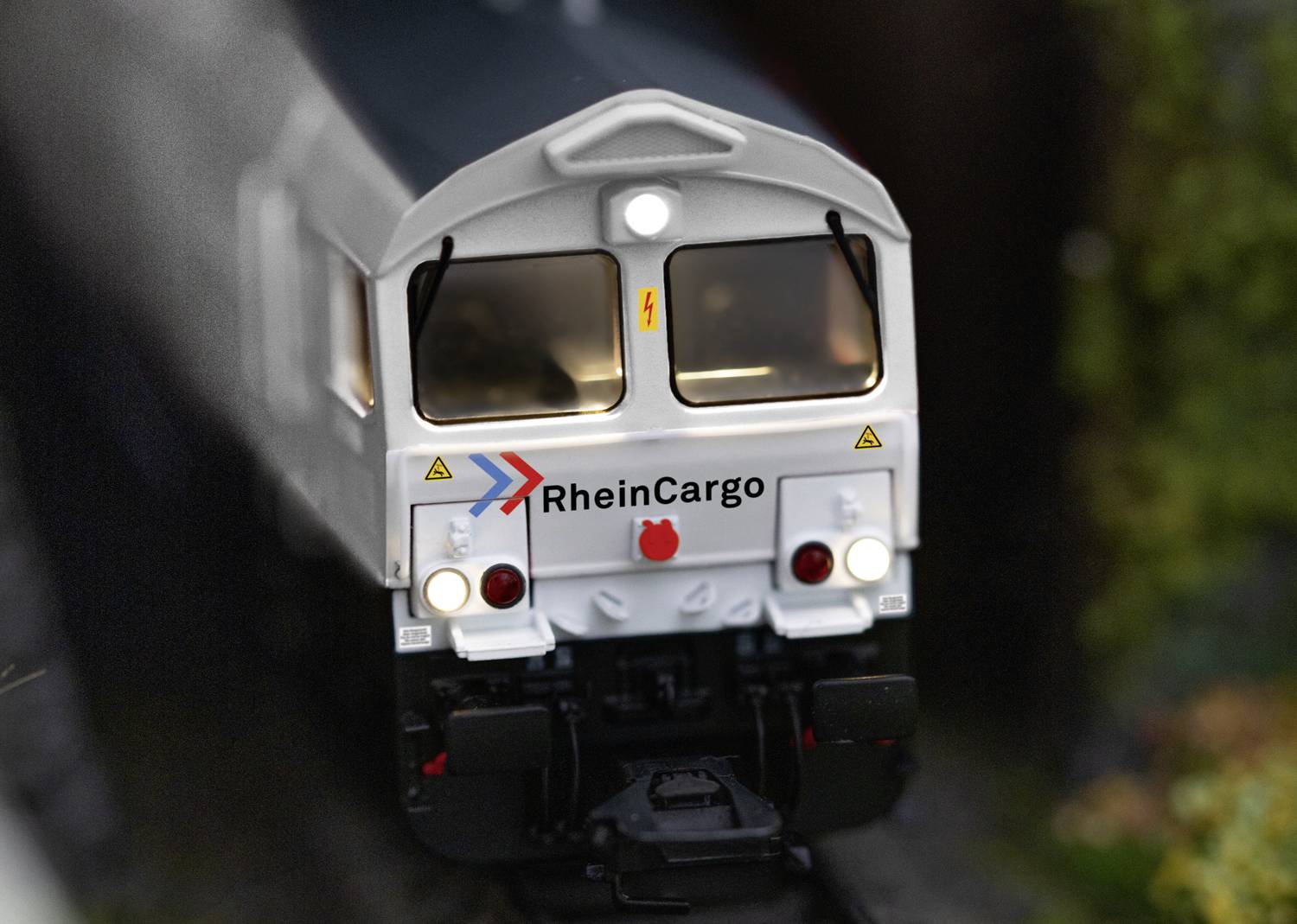 Ein Modellzug mit 'RheinCargo'-Logo fährt auf einer Bahnstrecke. Der Zug ist weiß mit grauen Details und befindet sich in einer landschaftlichen Umgebung.