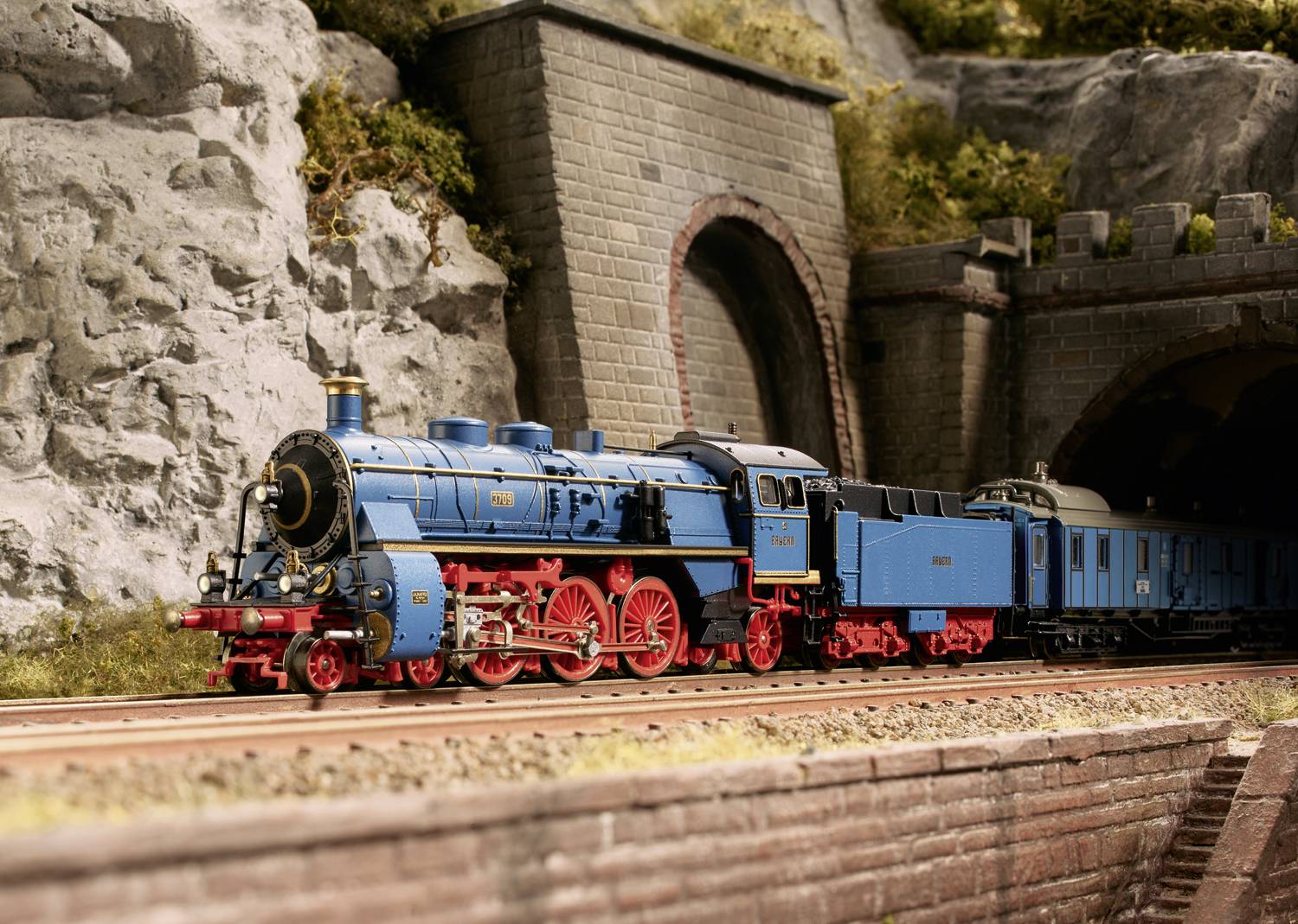 Ein blau-rote Dampflokomotive fährt aus einem Tunnel auf einer Modellbahnstrecke. Hintergrund mit Felsen und einem gemauerten Tunnel.
