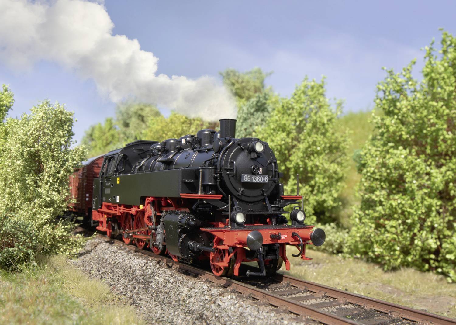 Eine schwarze Dampflokomotive fährt auf Schienen durch eine grüne Landschaft, umgeben von Bäumen und Büschen, mit Rauch aus dem Schornstein.