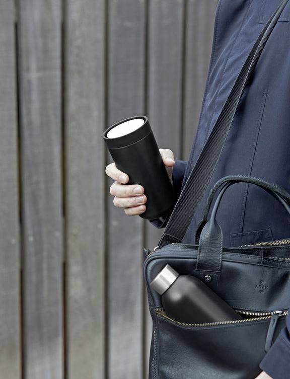 Eine Person hält einen schwarzen Becher und hat eine Tasche mit einer Flasche darin über der Schulter. Hintergrund: Holzwand.