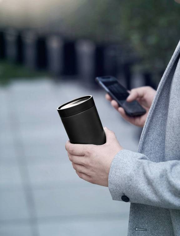 Person hält einen schwarzen Thermobecher in der linken Hand und ein Smartphone in der rechten Hand, während sie draußen spazieren geht.