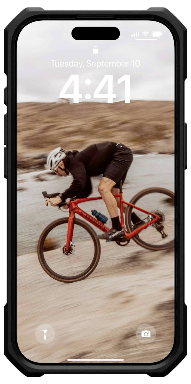 Eine Person fährt mit einem roten Fahrrad schnell einen Sandweg entlang. Hintergrund zeigt unscharfe Hügel und Wasser.