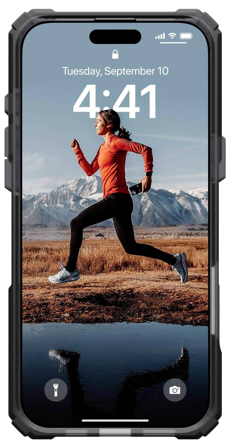 Eine Frau joggt in Sportkleidung auf einer Feldstraße vor schneebedeckten Bergen. Das Smartphone zeigt Uhrzeit und Datum an.