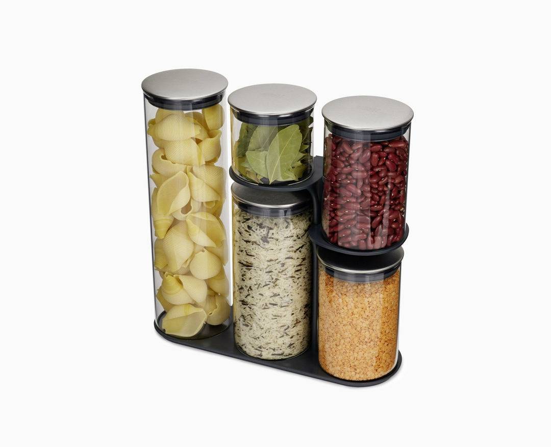 Drei hohe und zwei mittlere Glasbehälter, gefüllt mit Nudeln, Lorbeerblättern, roten Bohnen, Reis und roten Linsen, stehen auf einer schwarzen Ablage.
