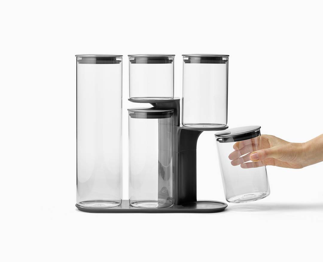 Eine Hand greift nach einem von fünf transparenten Vorratsgläsern, die auf einem schwarzen Gestell stehen.