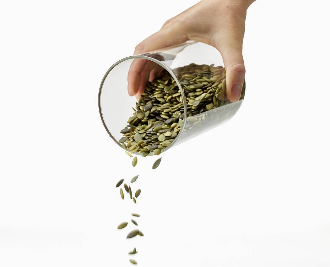 Eine Hand gießt Kürbiskerne aus einem Glas, die auf eine flache Oberfläche fallen. Die Kerne sind dunkelgrün und braun.