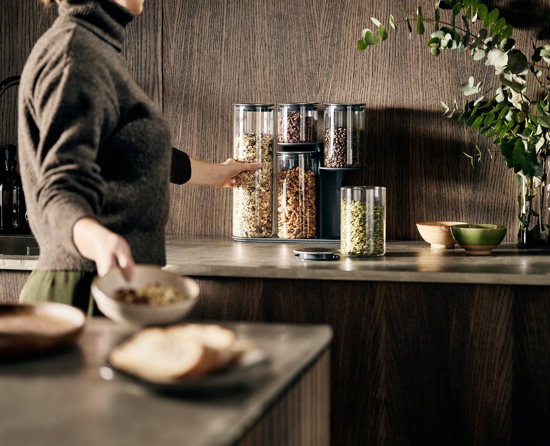 Eine Person bereitet in einer Küche Müsli vor, mit verschiedenen Behältern mit Körnern und Nüssen auf der Arbeitsplatte.