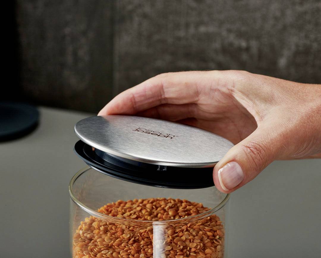 Hand öffnet Glas mit verschraubbarem Metalldeckel, voller roter Linsen, auf Küchenarbeitsplatte.