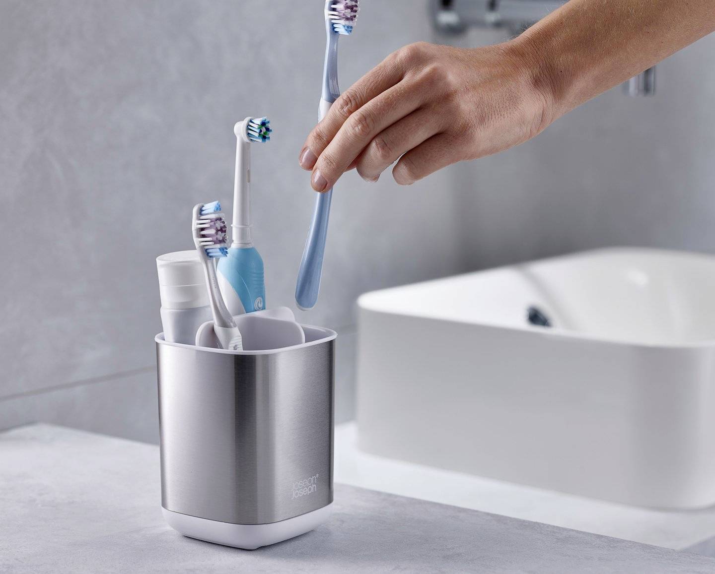 Eine Hand nimmt eine blaue Zahnbürste aus einem Edelstahlbecher, der auch andere Zahnbürsten und Zahnpasta enthält. Im Hintergrund ist ein Waschbecken zu sehen.