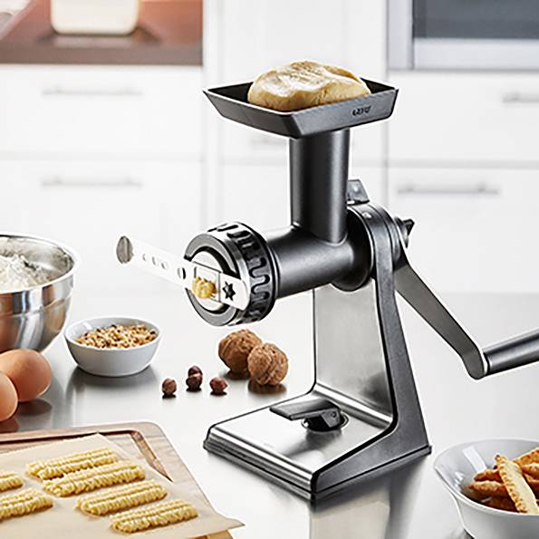 Handbetriebene Nudelmaschine in Küchenszene, Teig oben, fertige Nudeln unten. Weitere Zutaten: Mehl, Eier, Nüsse.