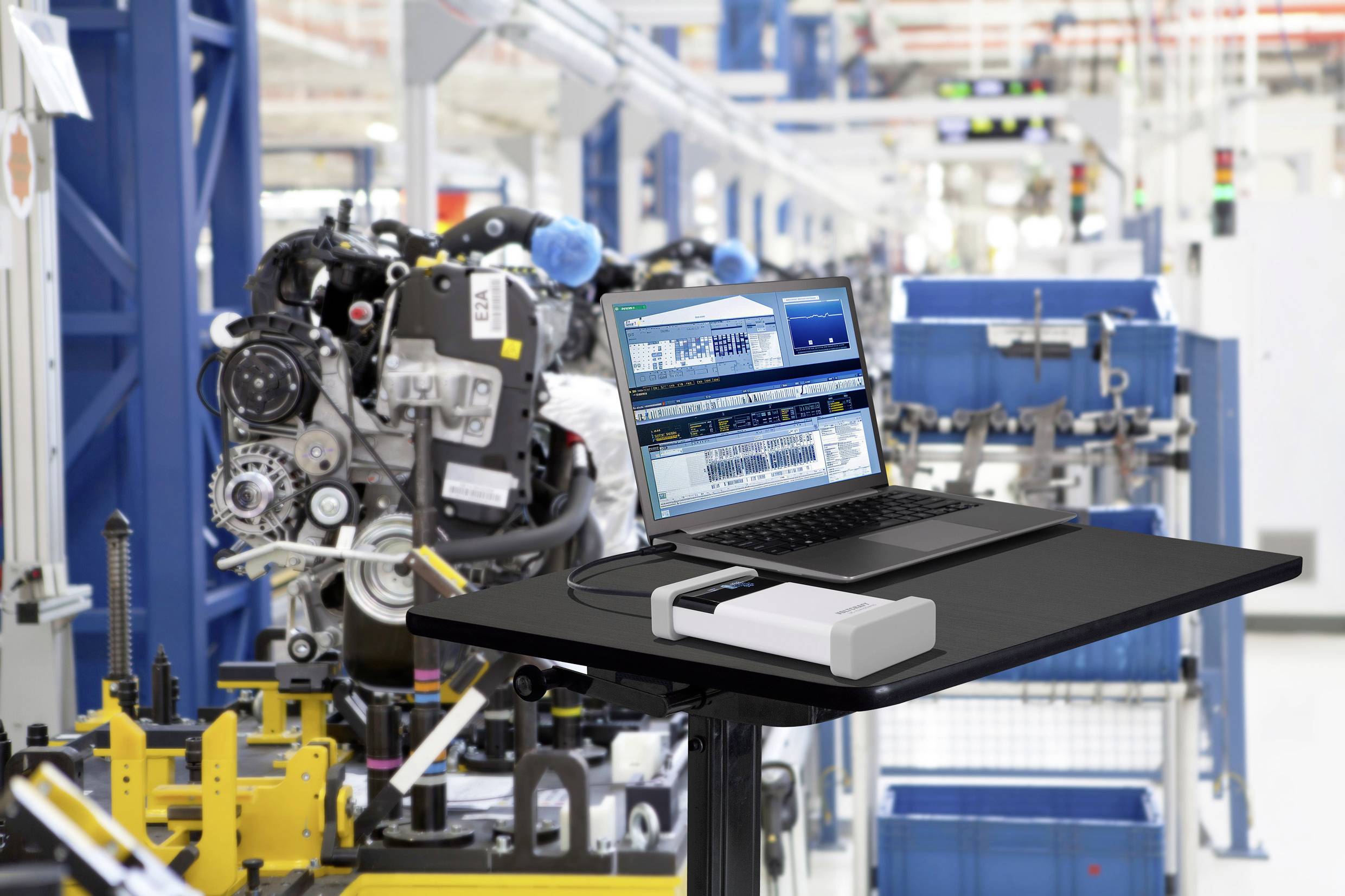 Ein Laptop steht auf einem Tisch in einer Fabrik. Der Bildschirm zeigt Fertigungssoftware. Im Hintergrund sind Maschinen und Produktionslinien zu sehen.