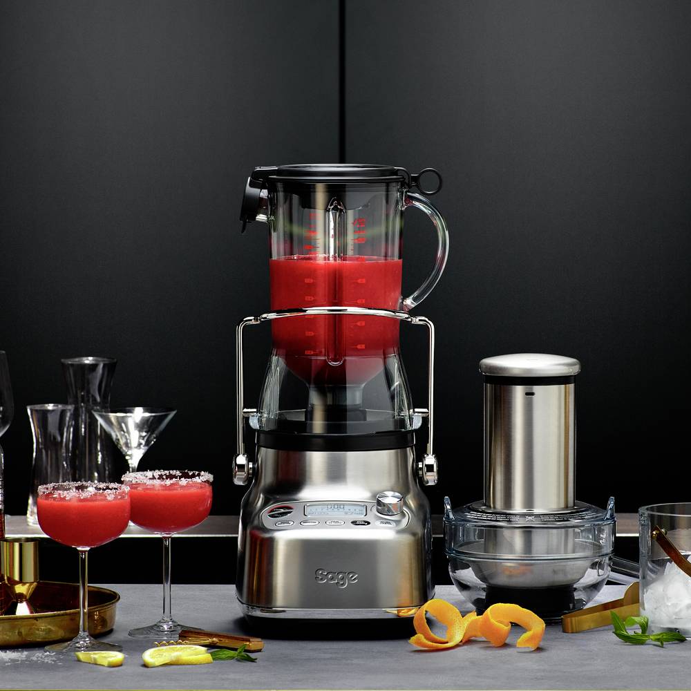 Ein Standmixer zerkleinert rote Früchte in einem modernen Küchenambiente. Daneben stehen Cocktailgläser, Orange und Limette.