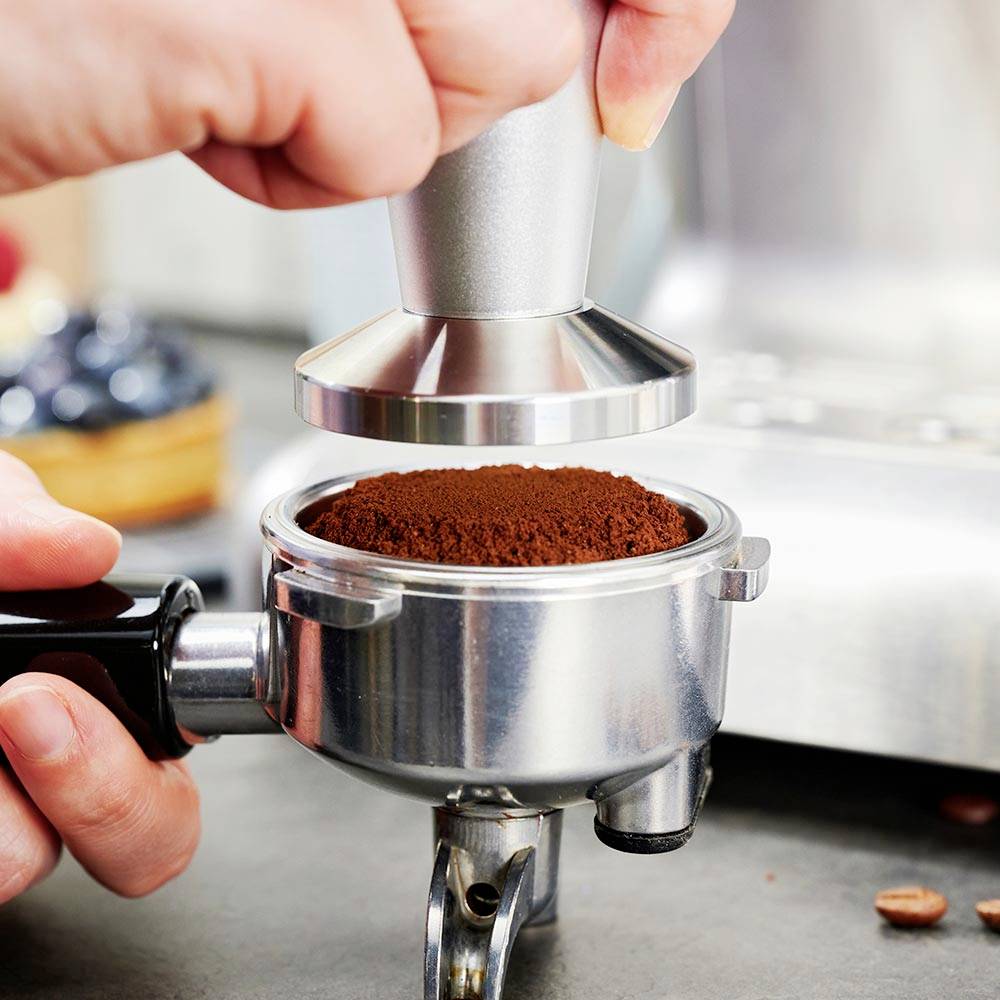Eine Hand presst Kaffeepulver mit einem Tamper in einen Siebträger, bereit für die Zubereitung von Espresso.