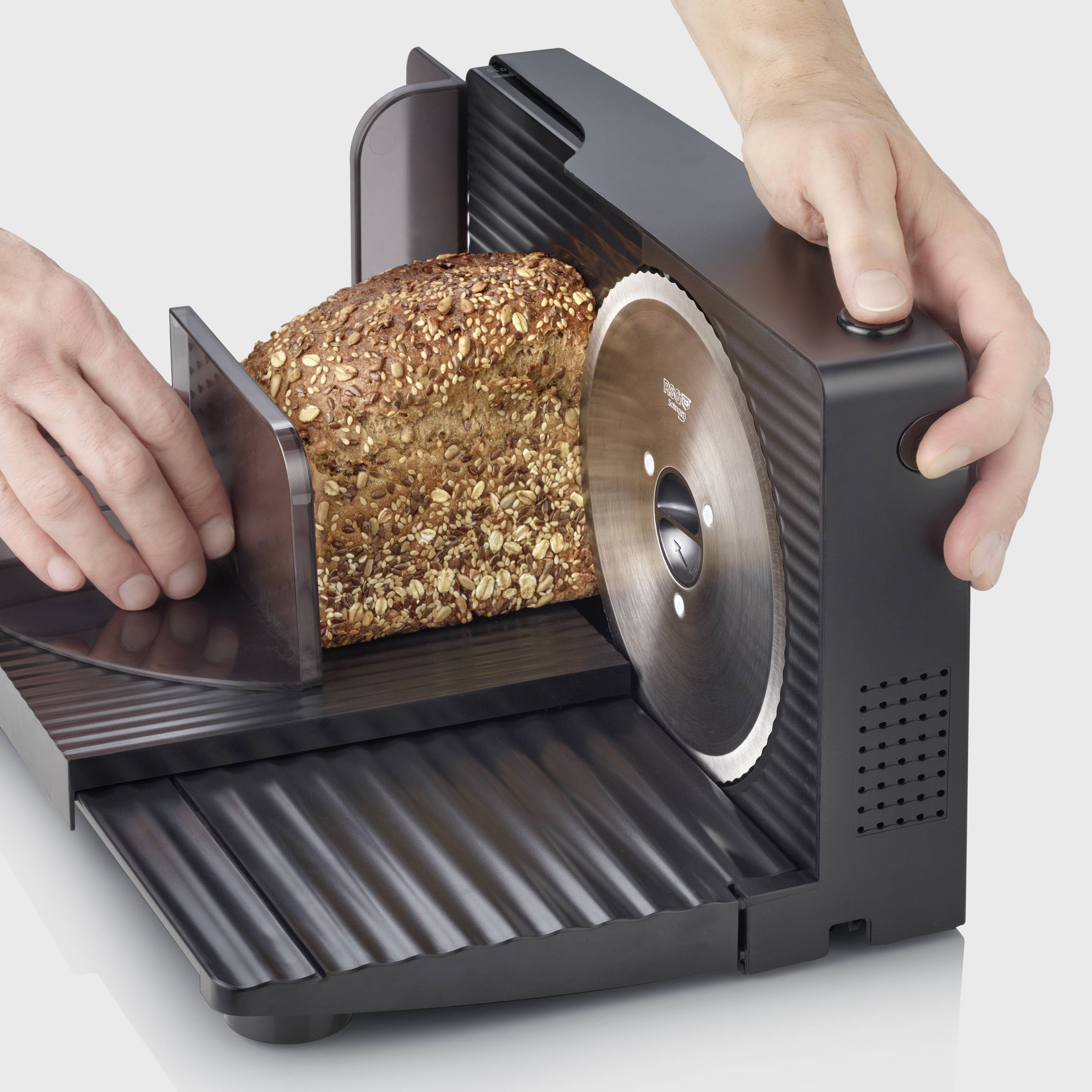 Ein Laib Vollkornbrot wird mit einer elektrischen Brotschneidemaschine geschnitten. Eine Hand führt das Brot an die Klinge.