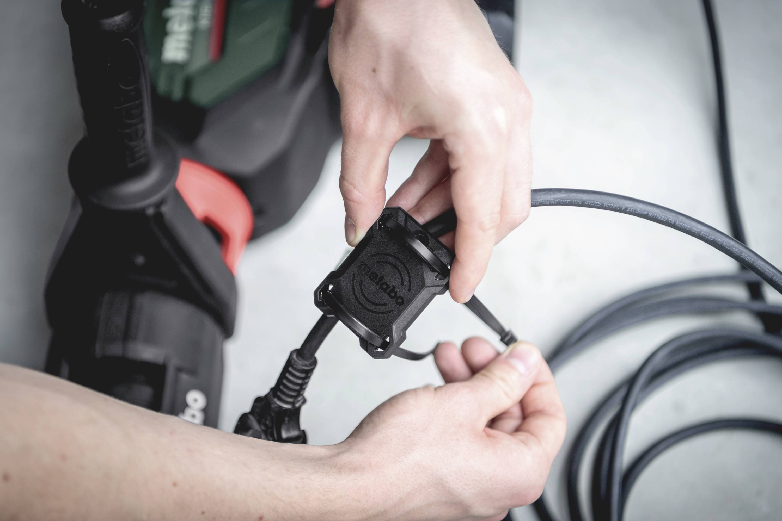Eine Person hält ein Elektrowerkzeug und verbindet ein Kabel. Hände justieren den Anschluss. Klarer Fokus auf die Verbindung und die Werkzeuge.