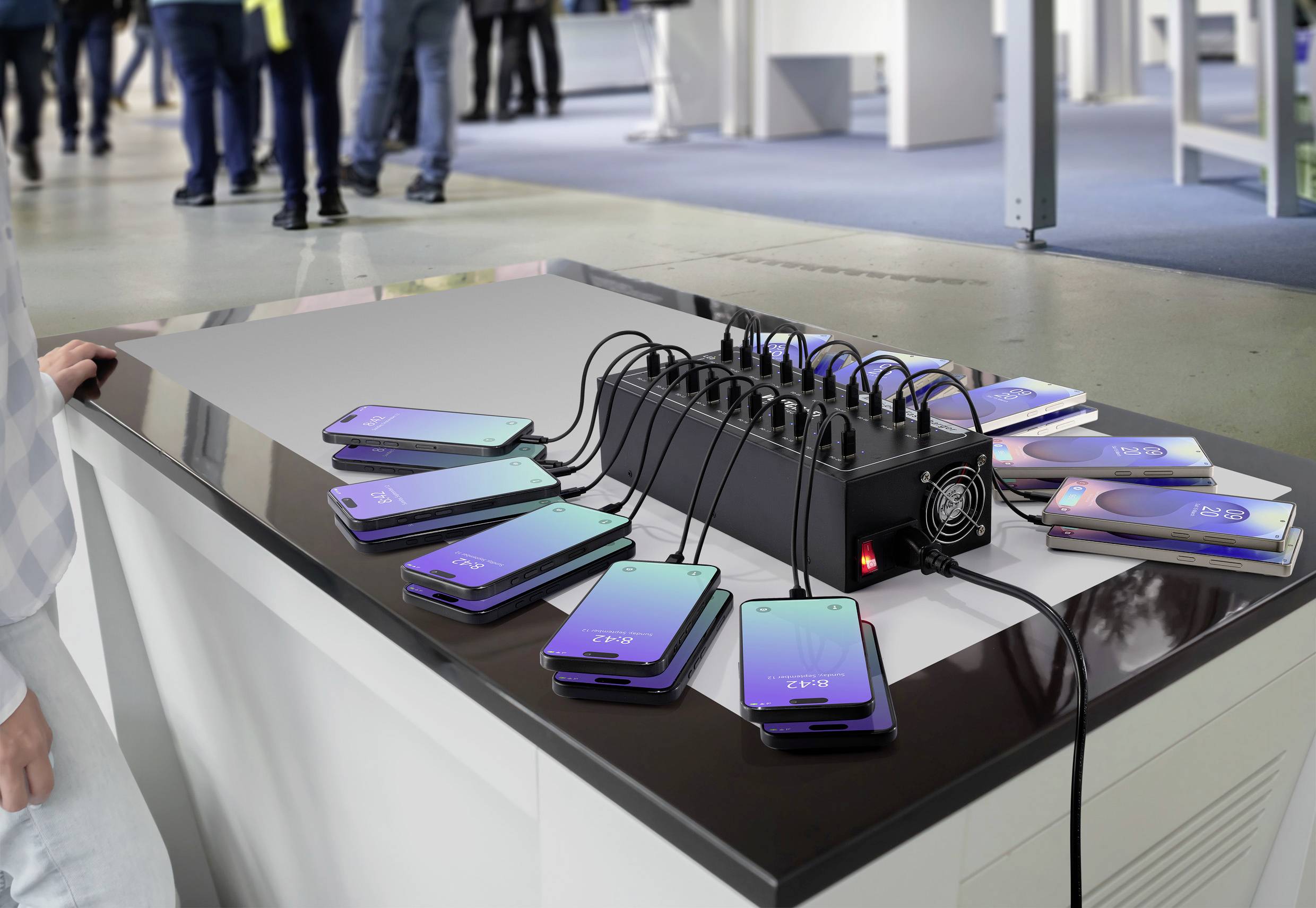 Mehrere Smartphones liegen auf einem Tisch und sind an eine zentrale Ladestation angeschlossen. Menschen gehen im Hintergrund vorbei.