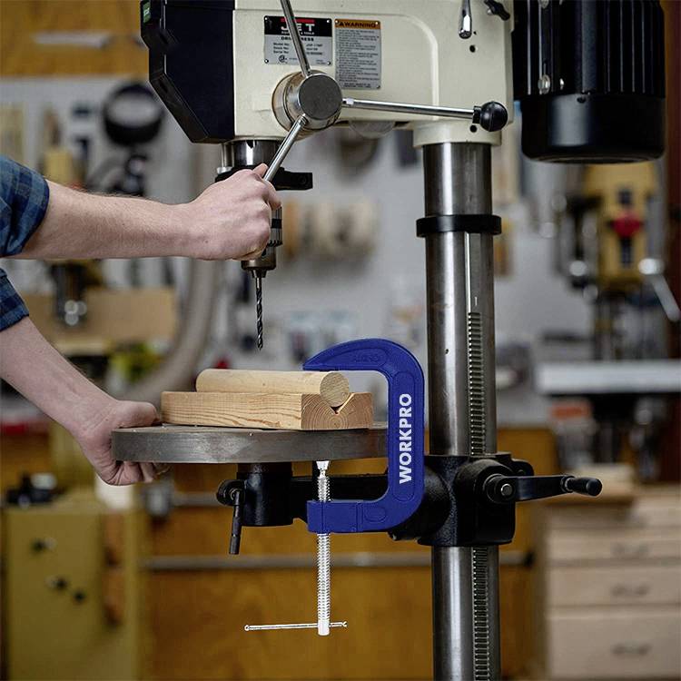 Eine Person bedient eine Tischbohrmaschine in einer Werkstatt. Ein Holzstück ist mit einer Klemme fixiert, während die Maschine ein Loch bohrt.