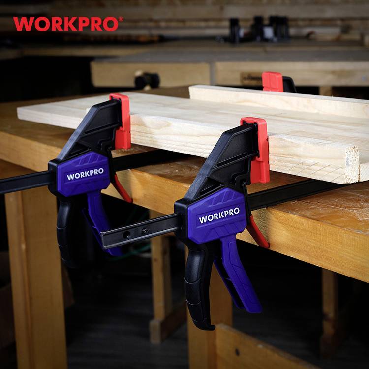 Holzwerkstatt mit zwei Werkstücken, die mit blauen und roten Klemmen an einem Tisch befestigt sind. Im Hintergrund ist unscharfes Werkzeug zu sehen.