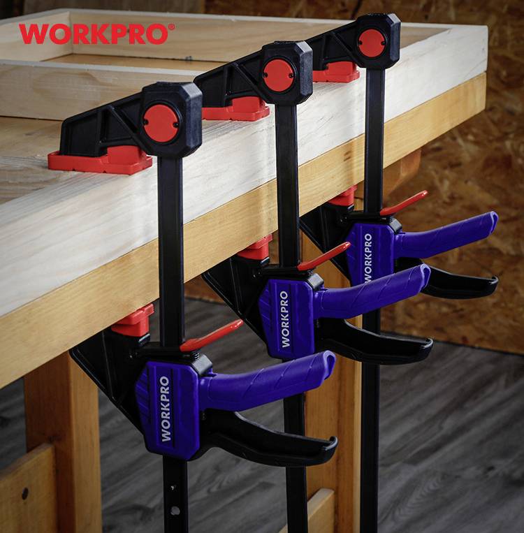 Drei blaue und schwarze Zwingen fixieren eine Holzkonstruktion auf einer Werkbank. Markenname 'WORKPRO' oben links in Rot.