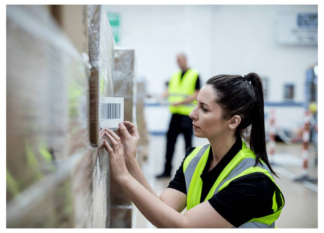Ein Arbeiter in einer Warnweste, konzentriert darauf, einen Barcode auf einer Palette in einem Lagerhaus zu scannen, während im Hintergrund ein weiterer Arbeiter steht.