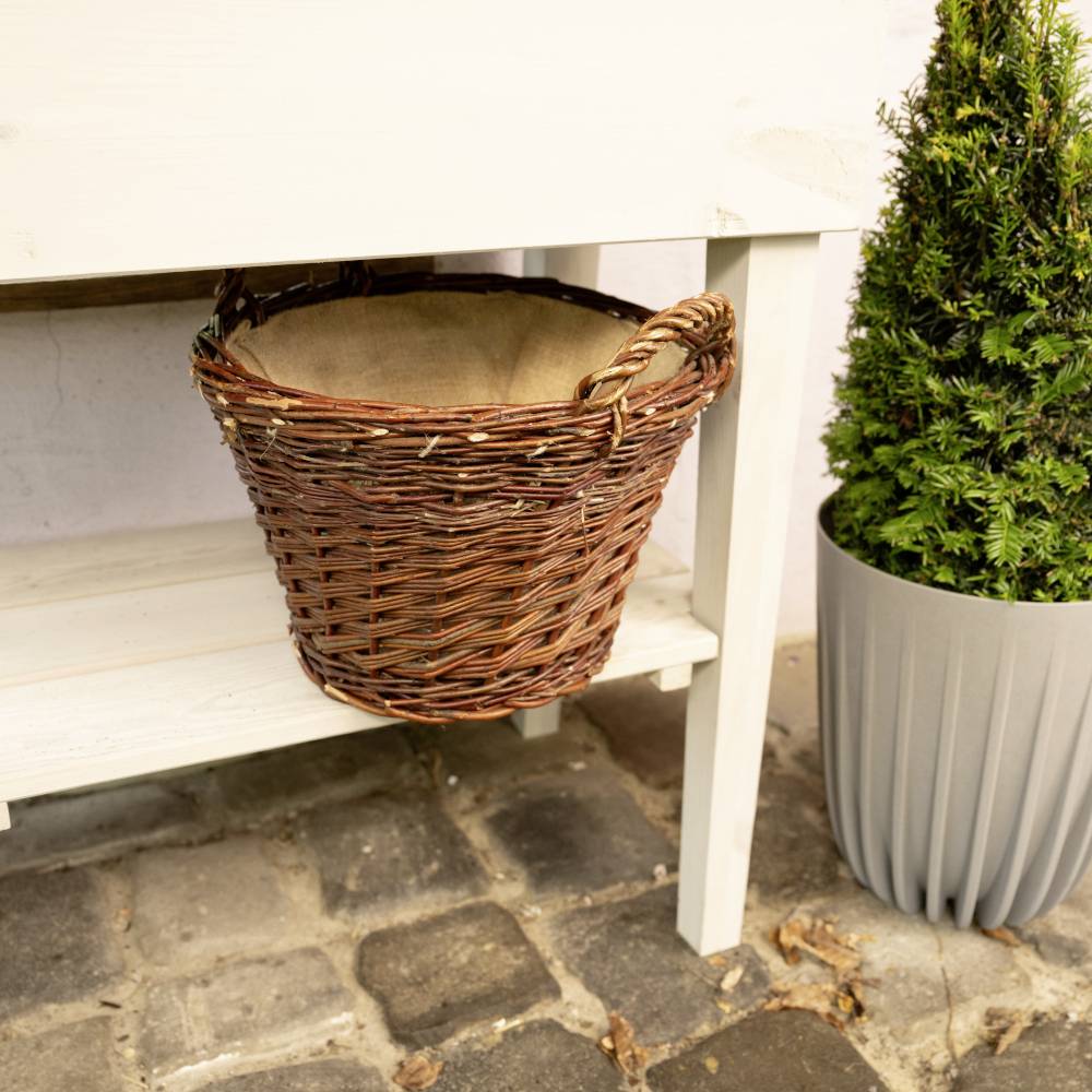 Ein leerer, brauner Weidenkorb steht auf einem weißen Regal im Außenbereich neben einer Topfpflanze auf einem gepflasterten Boden.