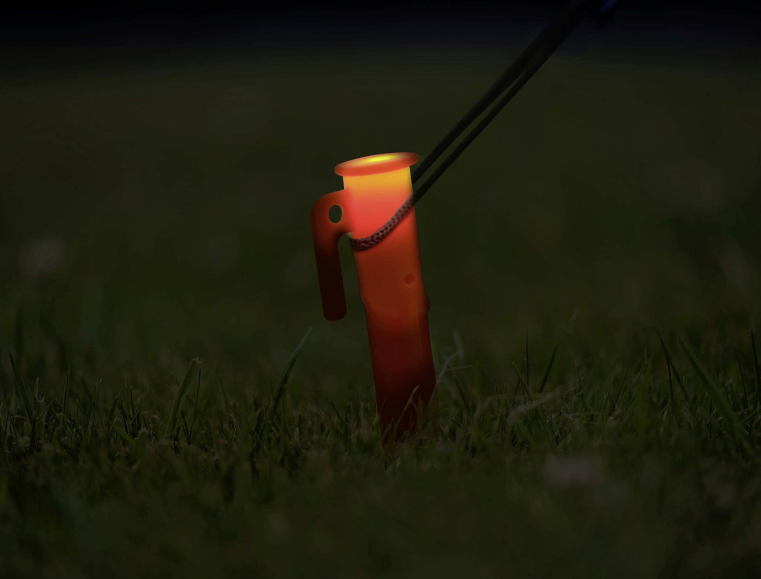 Eine leuchtende, orange Campinglaterne steht in einem dunklen Grasfeld, beleuchtet die Umgebung schwach.