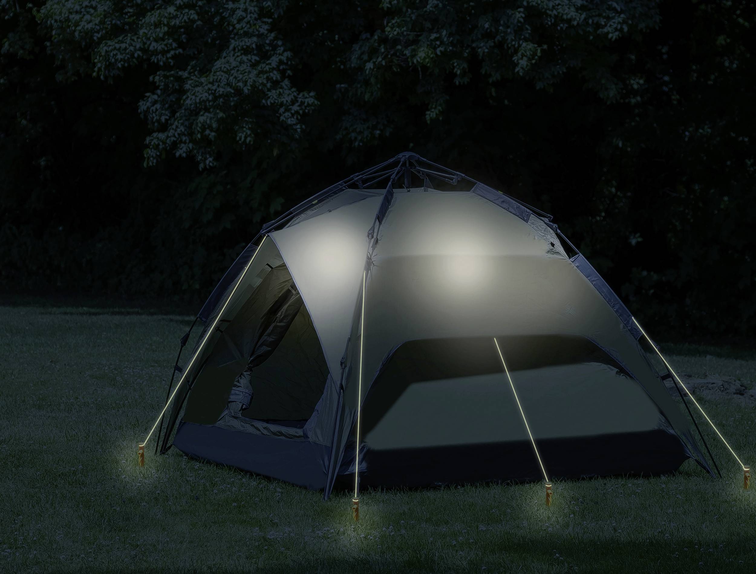 Ein Zelt steht nachts auf einer Wiese. Es ist beleuchtet und von Bäumen umgeben.