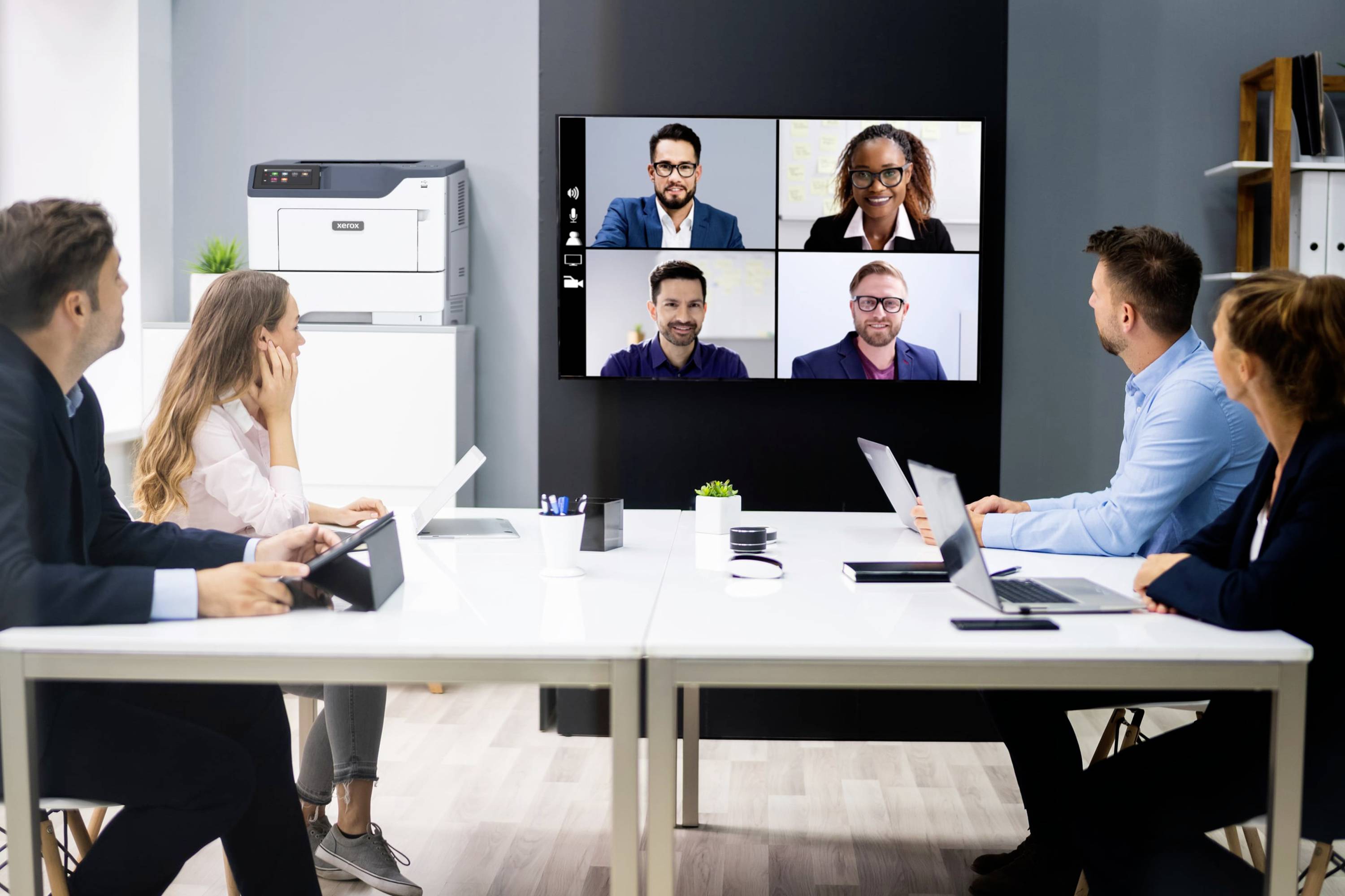 Sechs Personen in einem Konferenzraum nehmen an einem virtuellen Meeting teil. Vier Personen sind auf einem Bildschirm an der Wand zu sehen.