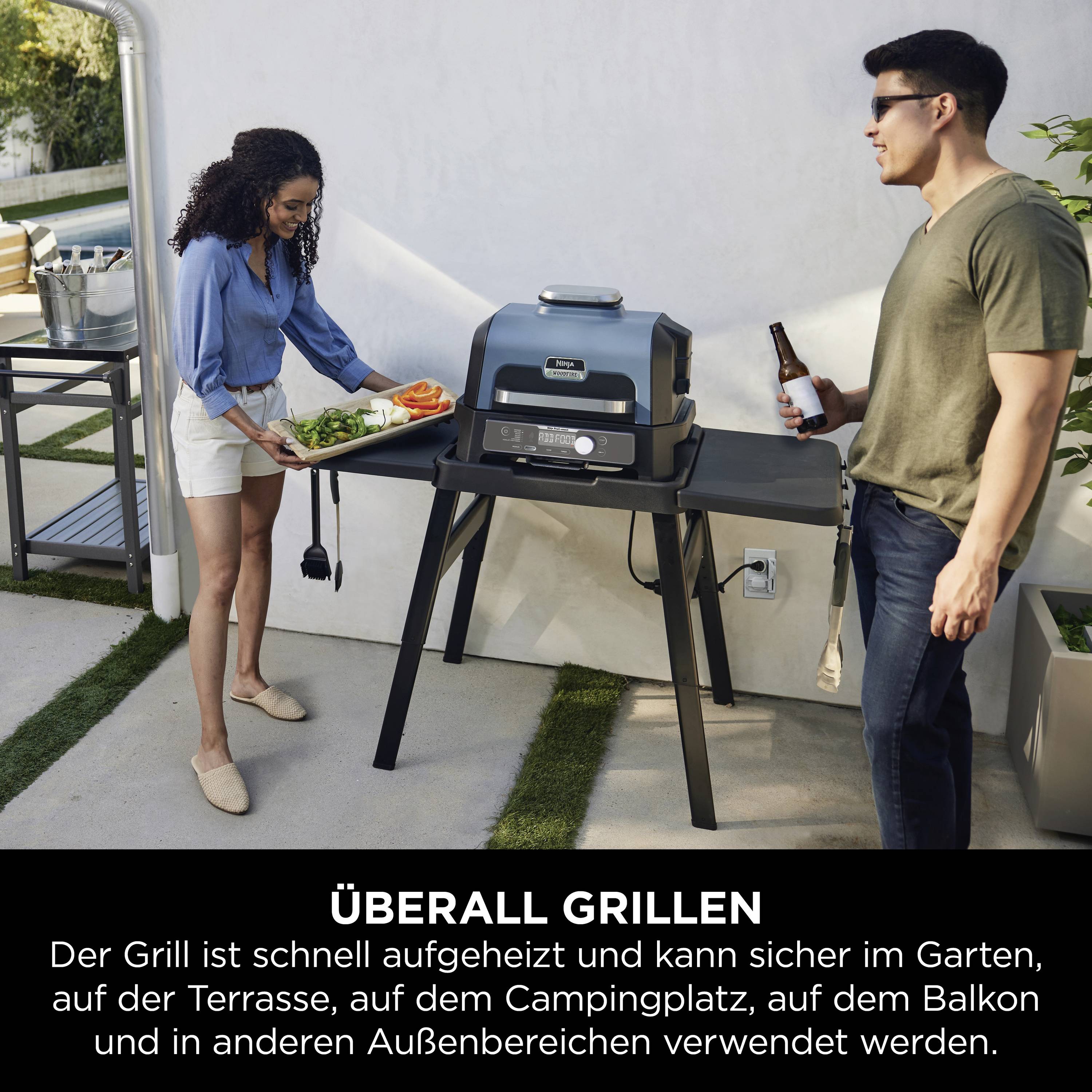 Eine Frau bringt Gemüse zu einem Grill, ein Mann hält ein Getränk. Der Grill steht im Garten neben einem Tisch mit Utensilien.