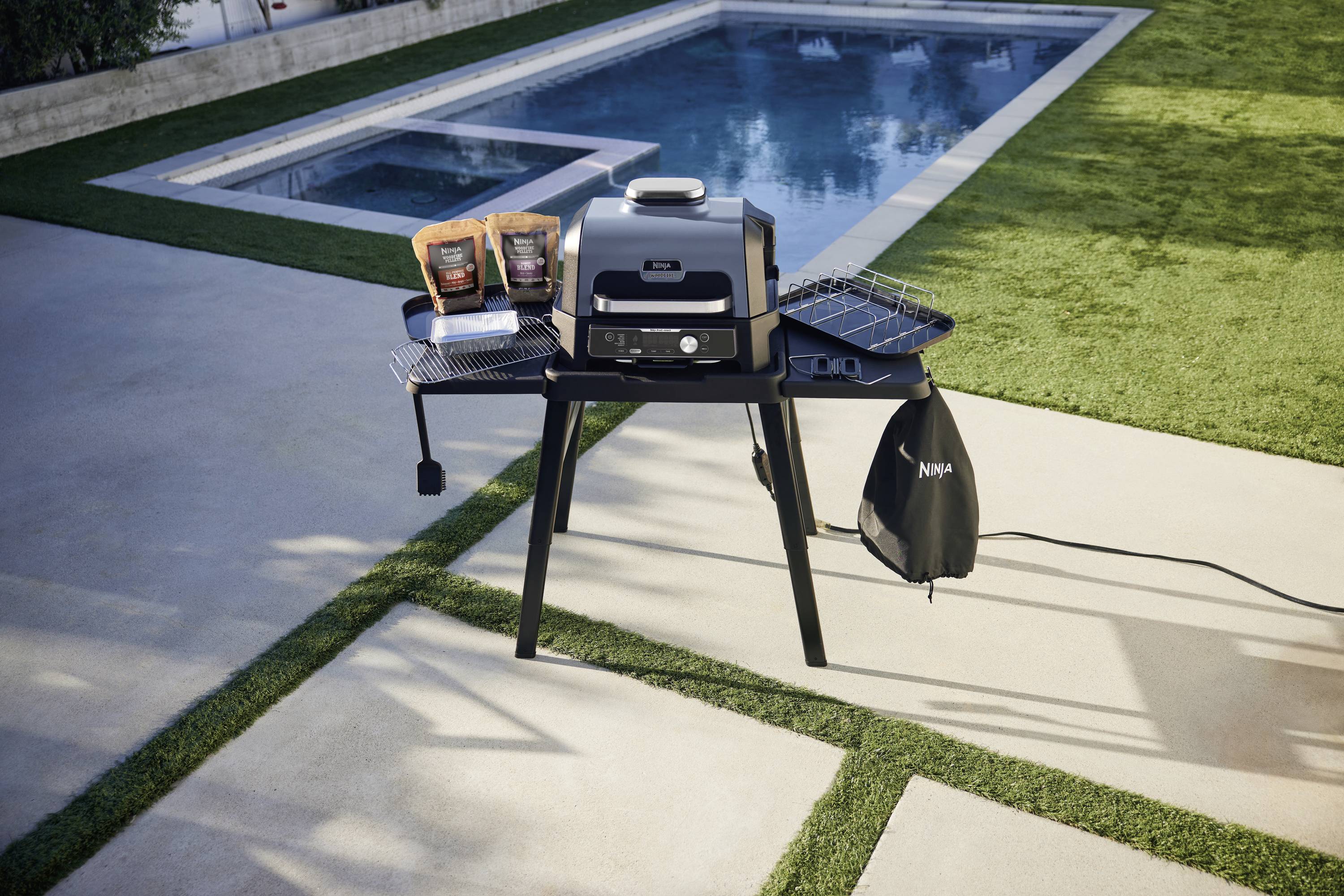 Gasgrill im Garten neben einem Pool, mit zwei Packungen Chips und einer Grillabdeckung auf den Ablageflächen. Sonniges Wetter.