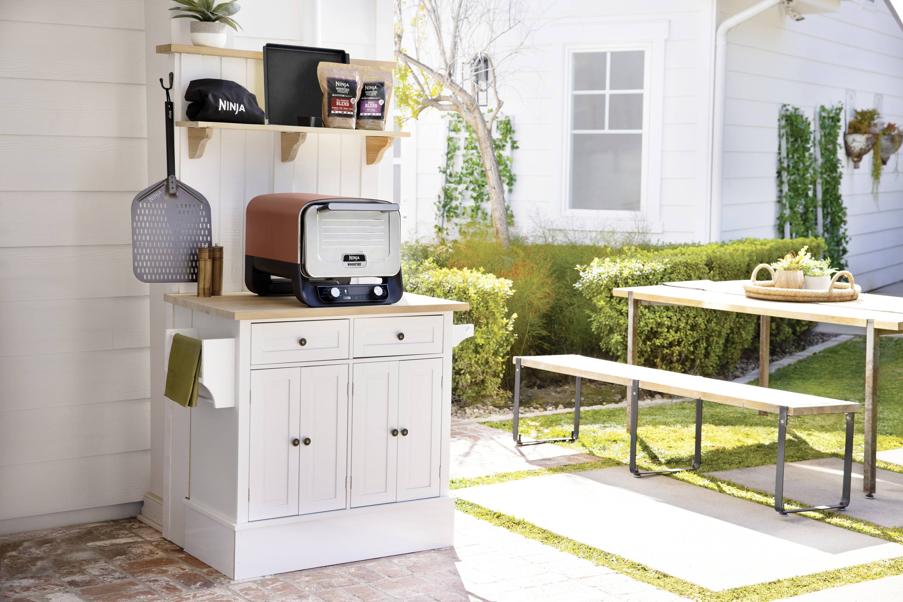 Eine Outdoor-Kochstation mit einem Ofen, Küchenutensilien und einem Grillplatz. Ein Tisch und Bänke stehen neben einem Garten.