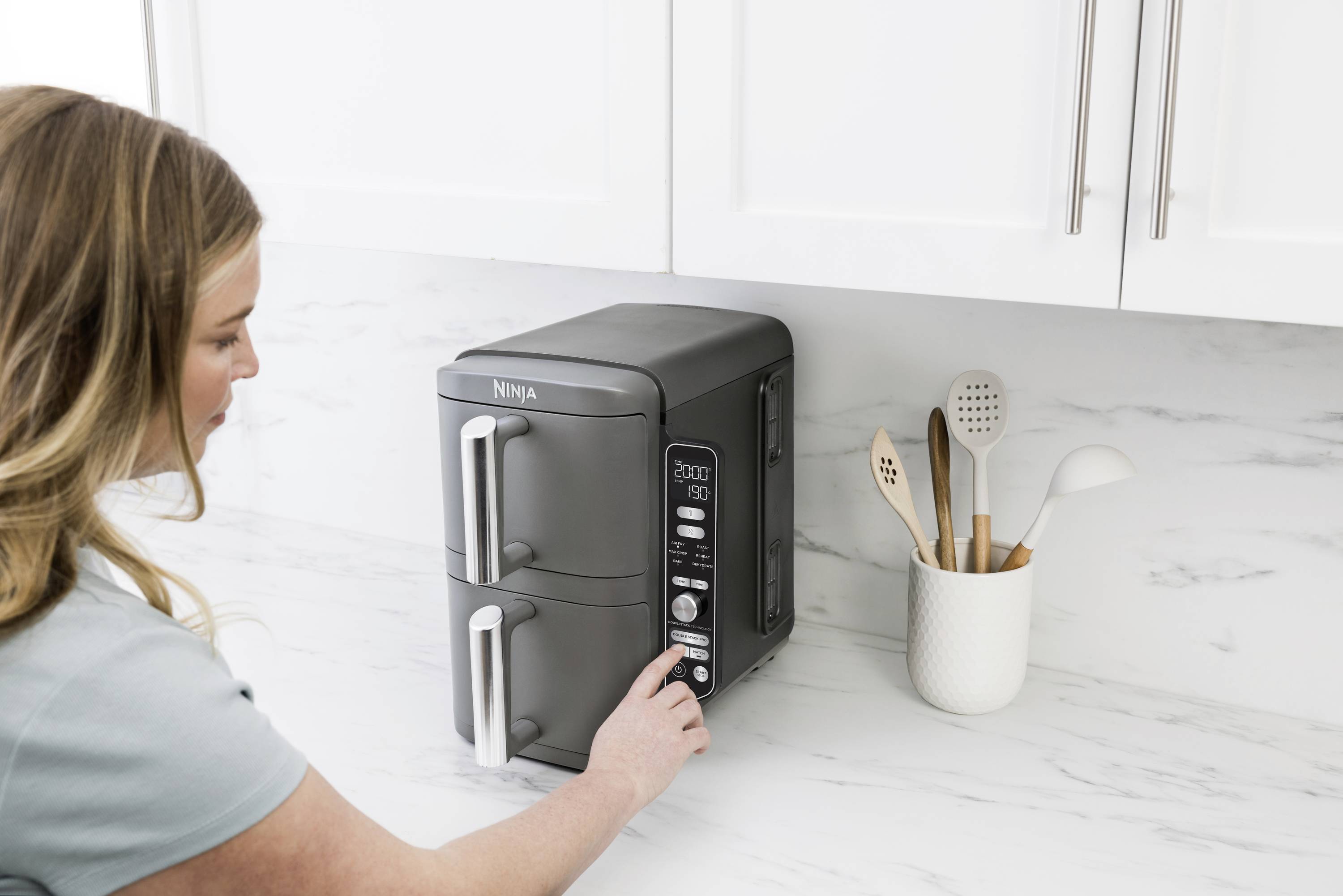 Eine Frau bedient einen schwarzen Doppelschubladen-Toasterofen in einer weißen Küche, daneben ein Behälter mit Kochutensilien.