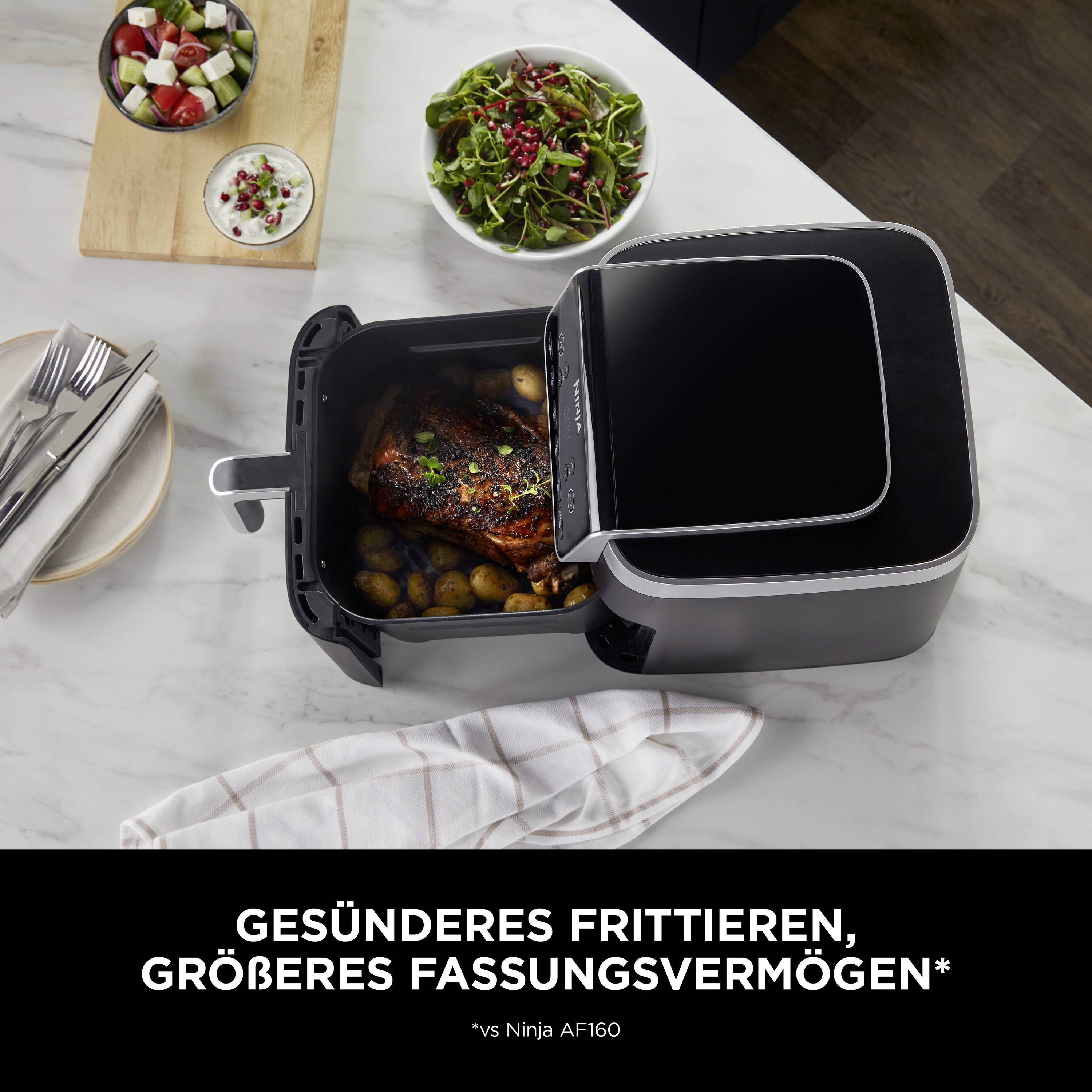 Luftfritteuse auf Küchentisch mit Hähnchen und Kartoffeln, umgeben von Beilagen in Schalen. Text: Gesünderes Frittieren, größeres Fassungsvermögen.