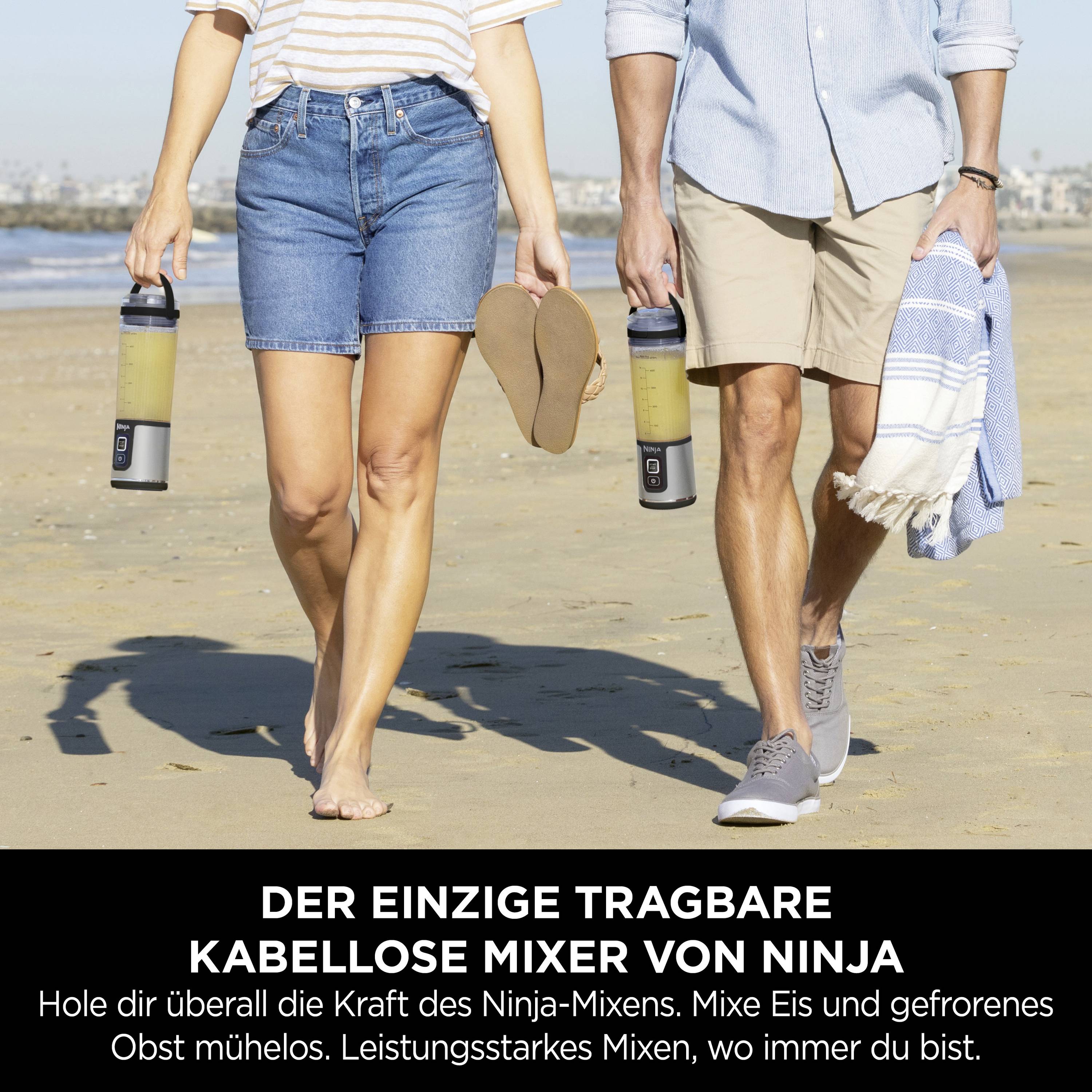 Zwei Personen gehen am Strand, beide halten tragbare Ninja-Mixer und gefrorene Getränke, Sonne und Stadt im Hintergrund.