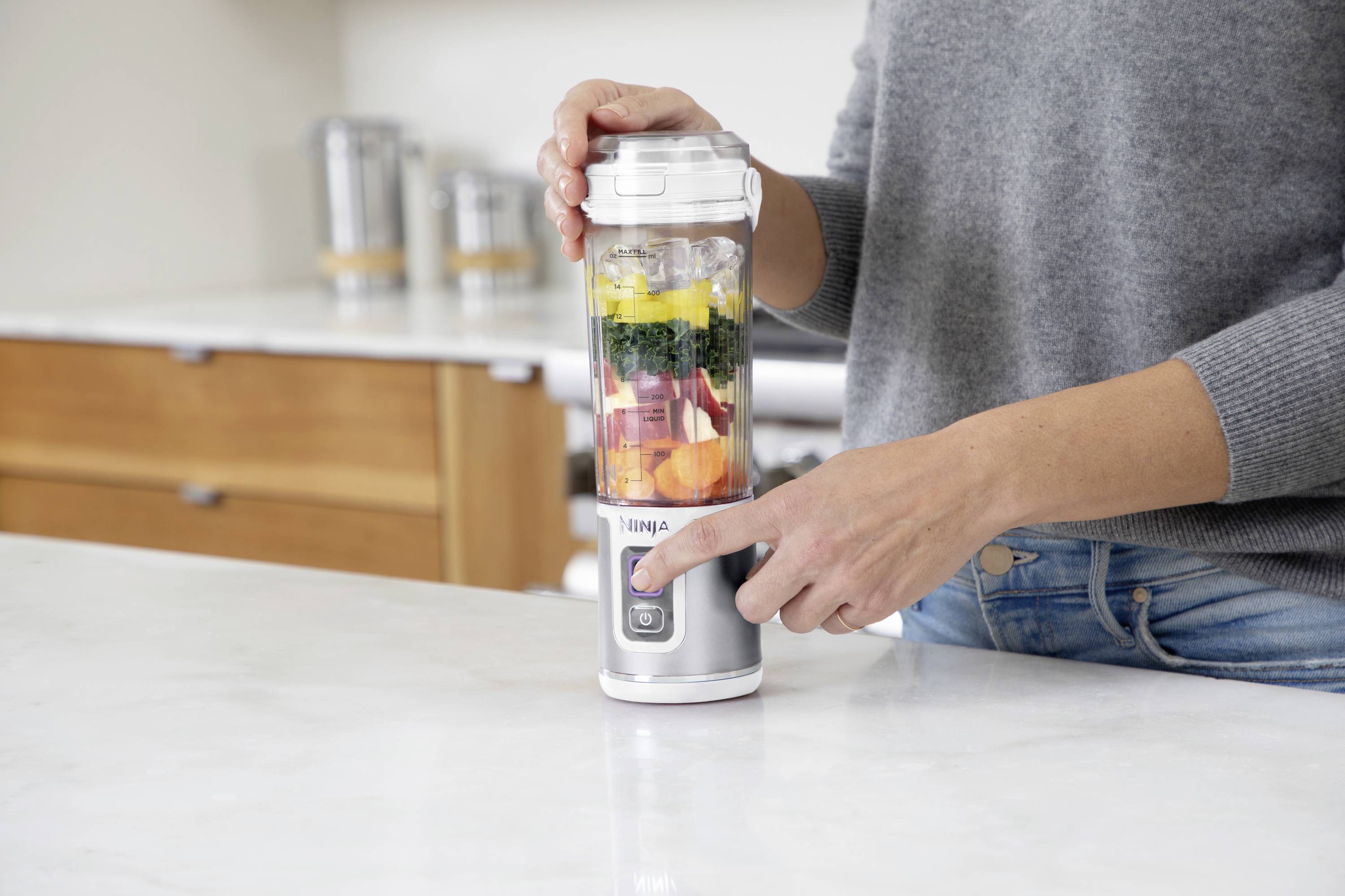 Eine Person bedient einen Standmixer mit verschiedenen bunten Zutaten, die Schichten von Obst und Gemüse im Glas zeigen.