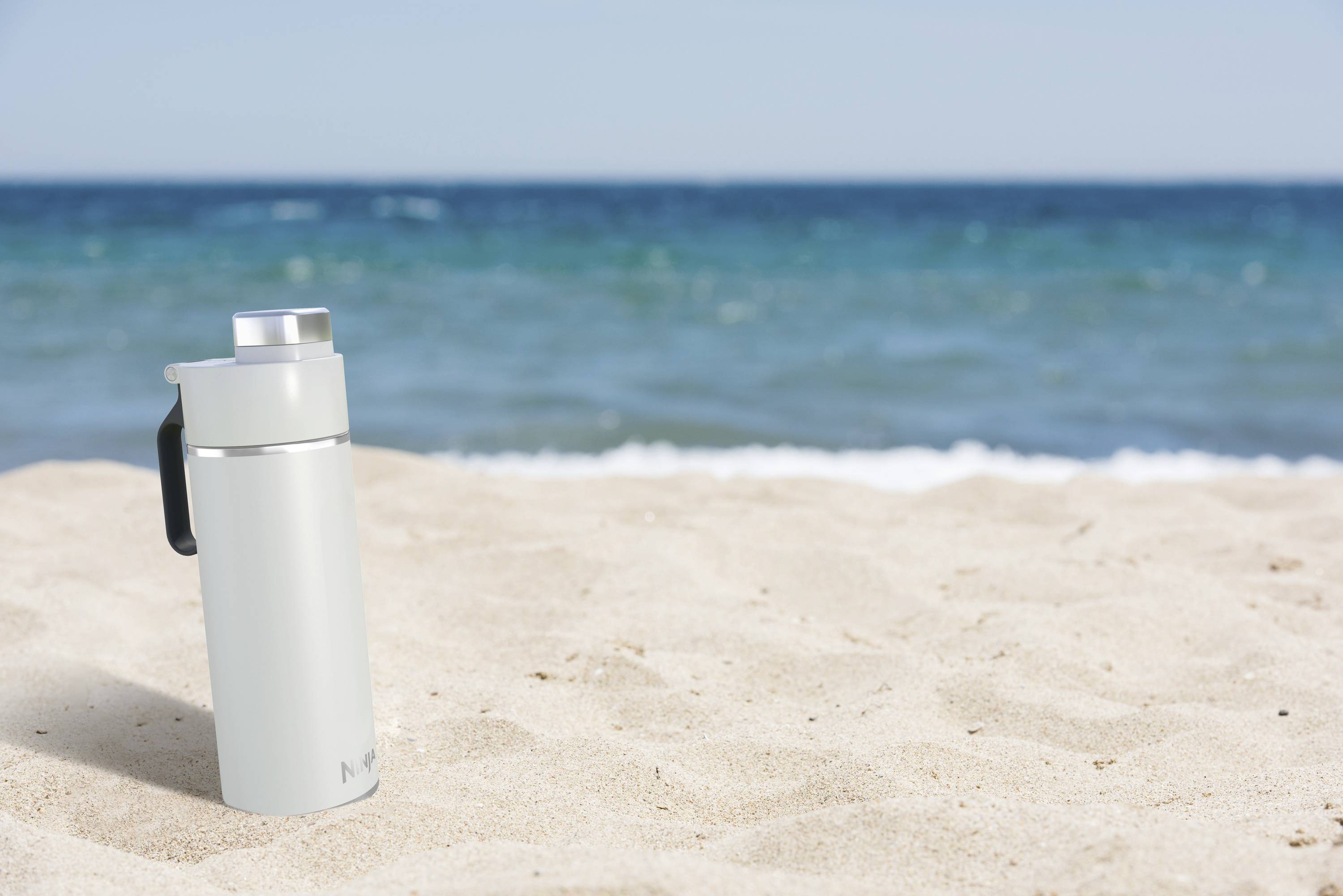 Ein silberner Thermobecher steht auf hellem Sand vor einem blauen Meer. Der Himmel ist klar.