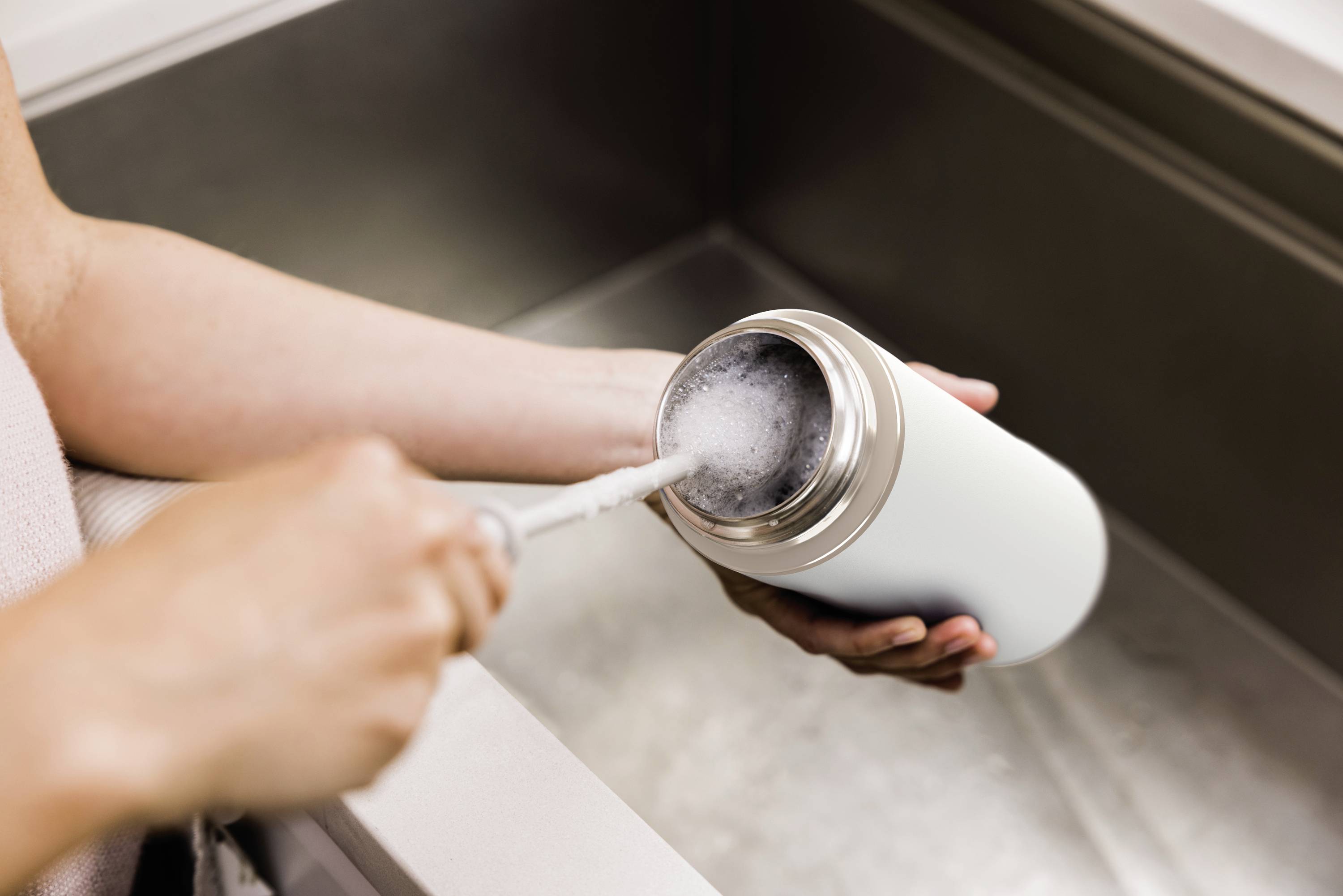 Eine Person reinigt eine weiße Wasserflasche mit einer Bürste über einem Waschbecken.
