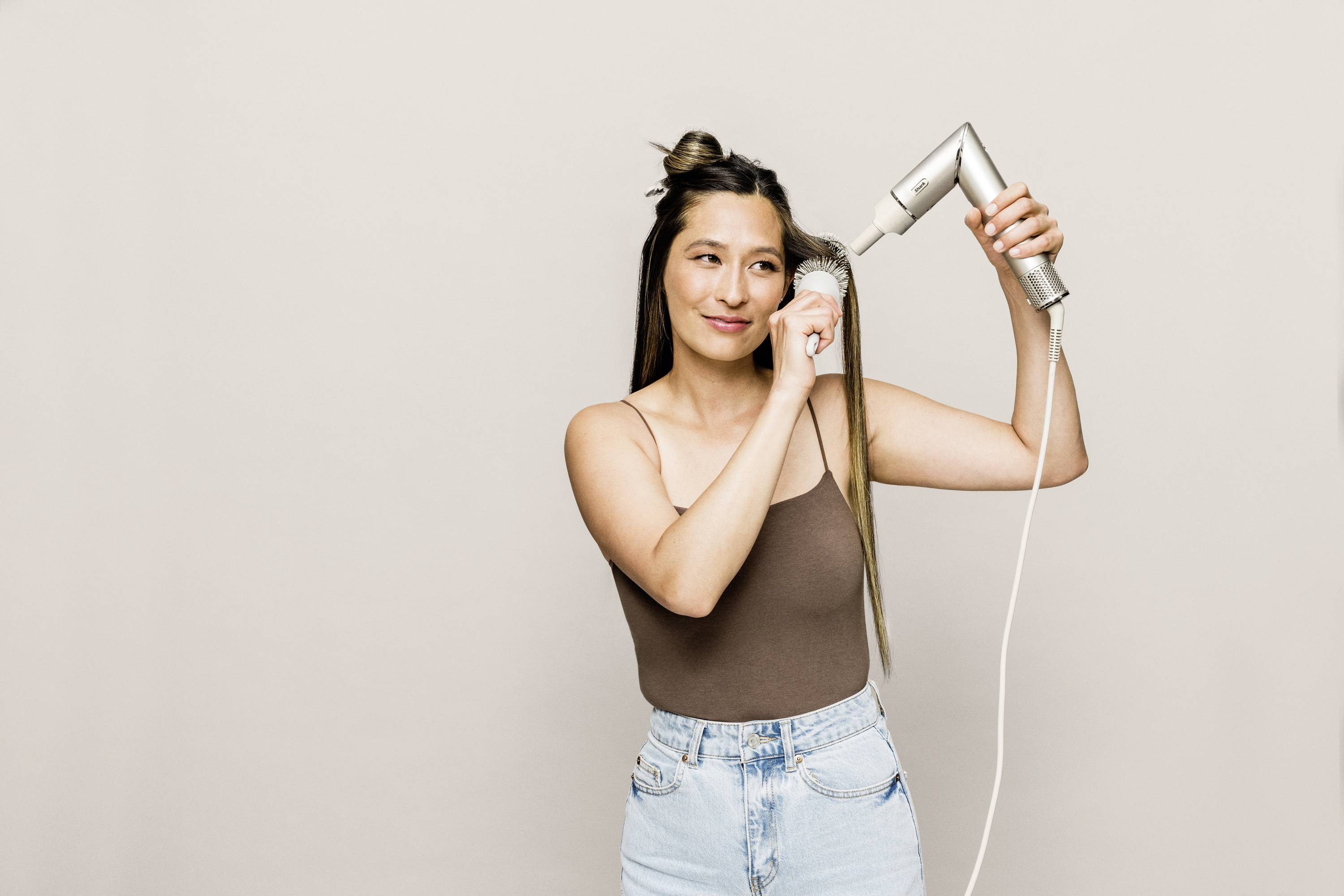 Frau trocknet ihr langes Haar mit einem Haartrockner, trägt ein braunes Top und Jeans, lächelt leicht, vor neutralem Hintergrund.