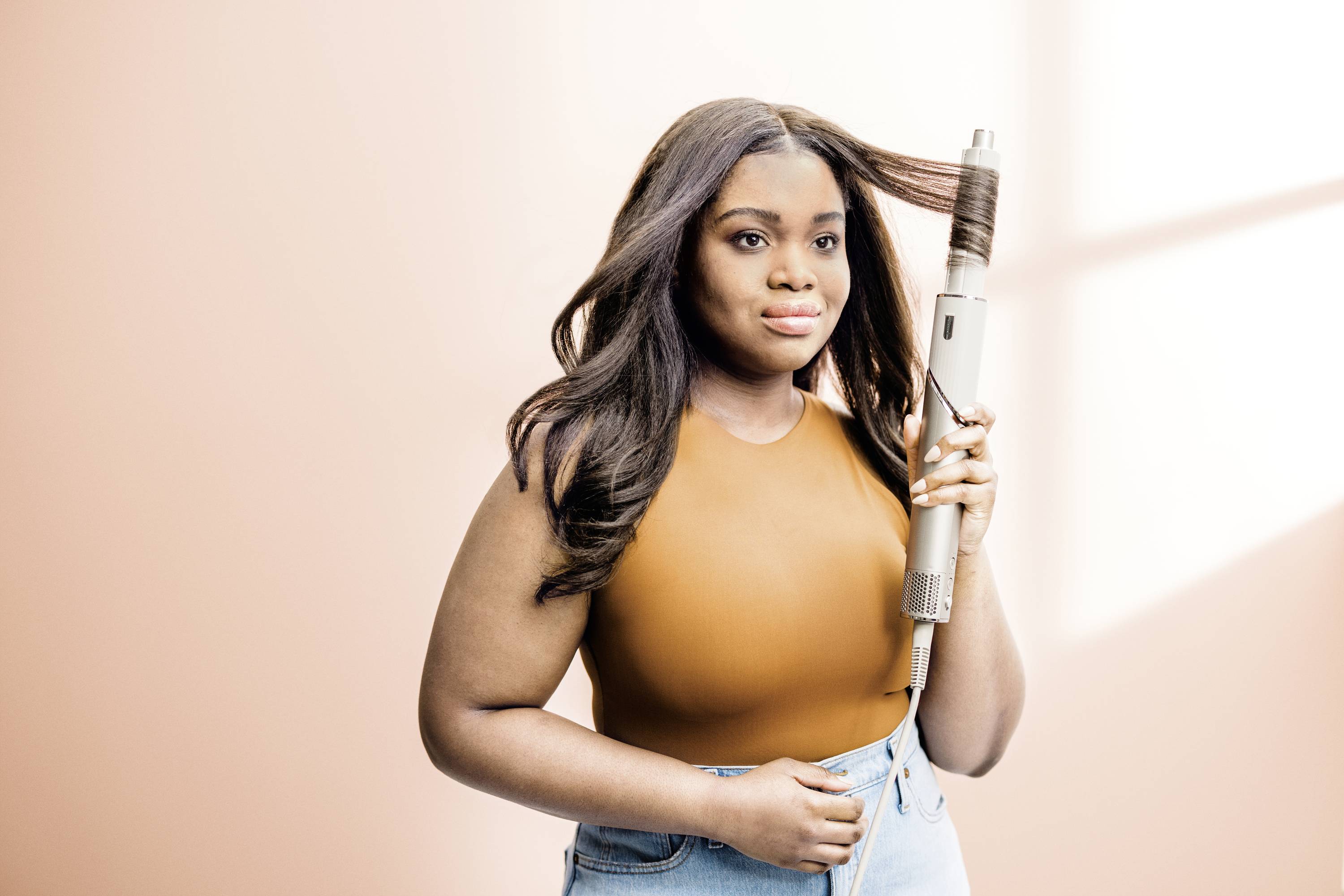 Eine Frau mit langen Haaren verwendet einen Lockenstab. Sie trägt ein oranges Oberteil und Jeans, im Hintergrund ist eine helle Wand.