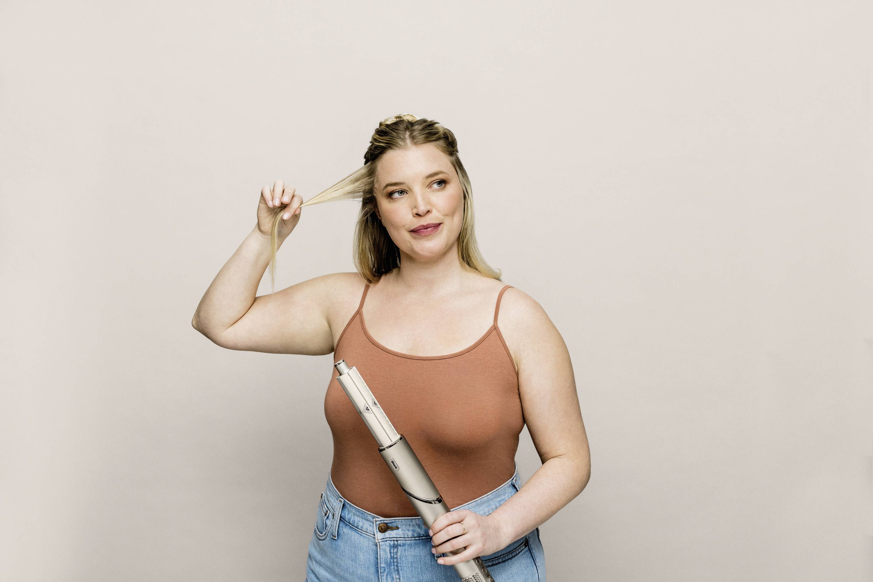 Frau mit blonden Haaren hält eine Haarsträhne und einen Lockenstab. Sie trägt ein braunes Top und blaue Jeans vor neutralem Hintergrund.