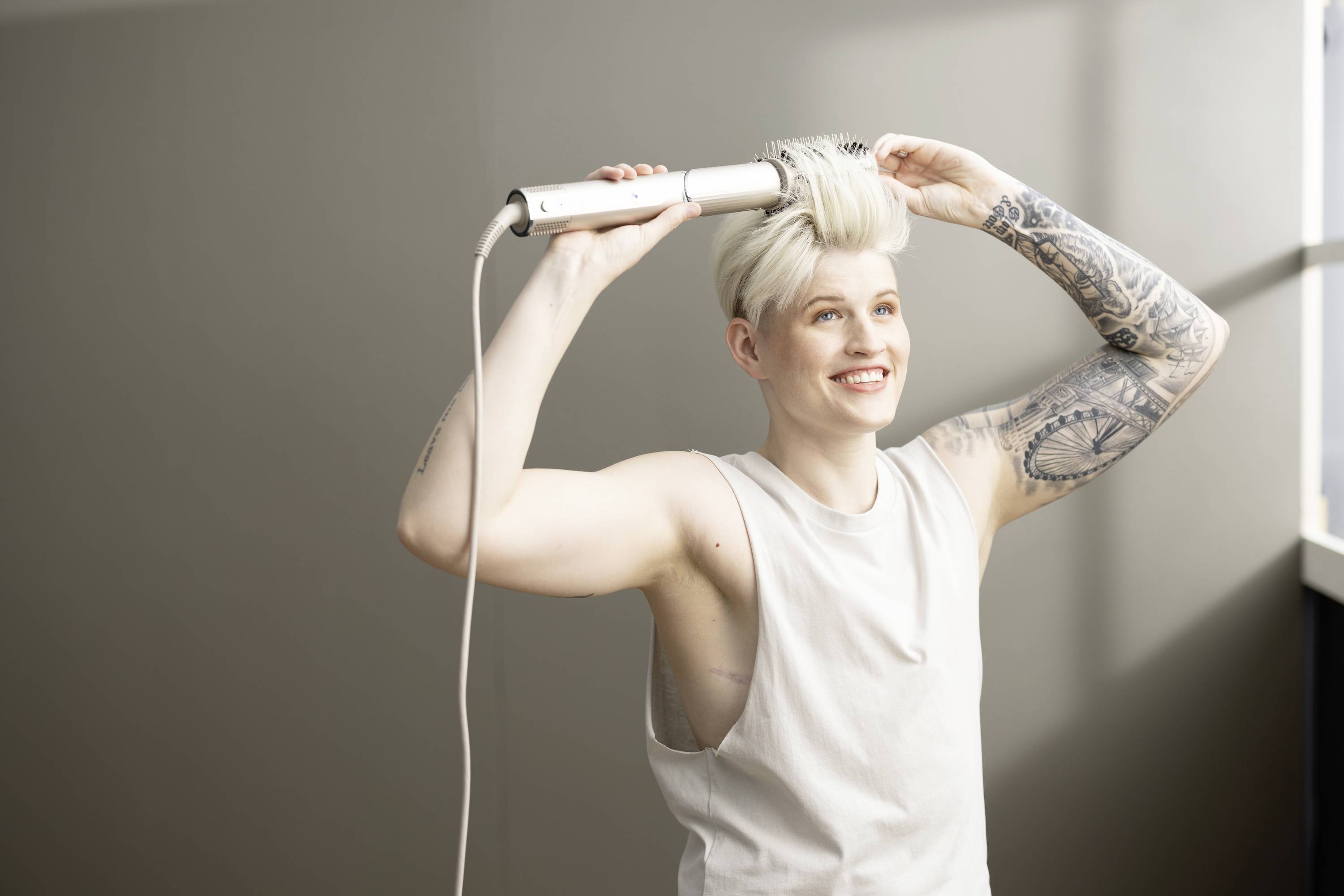 Eine Person mit Tattoos föhnt ihr blondes, kurzes Haar mit einer Rundbürste und lächelt dabei.