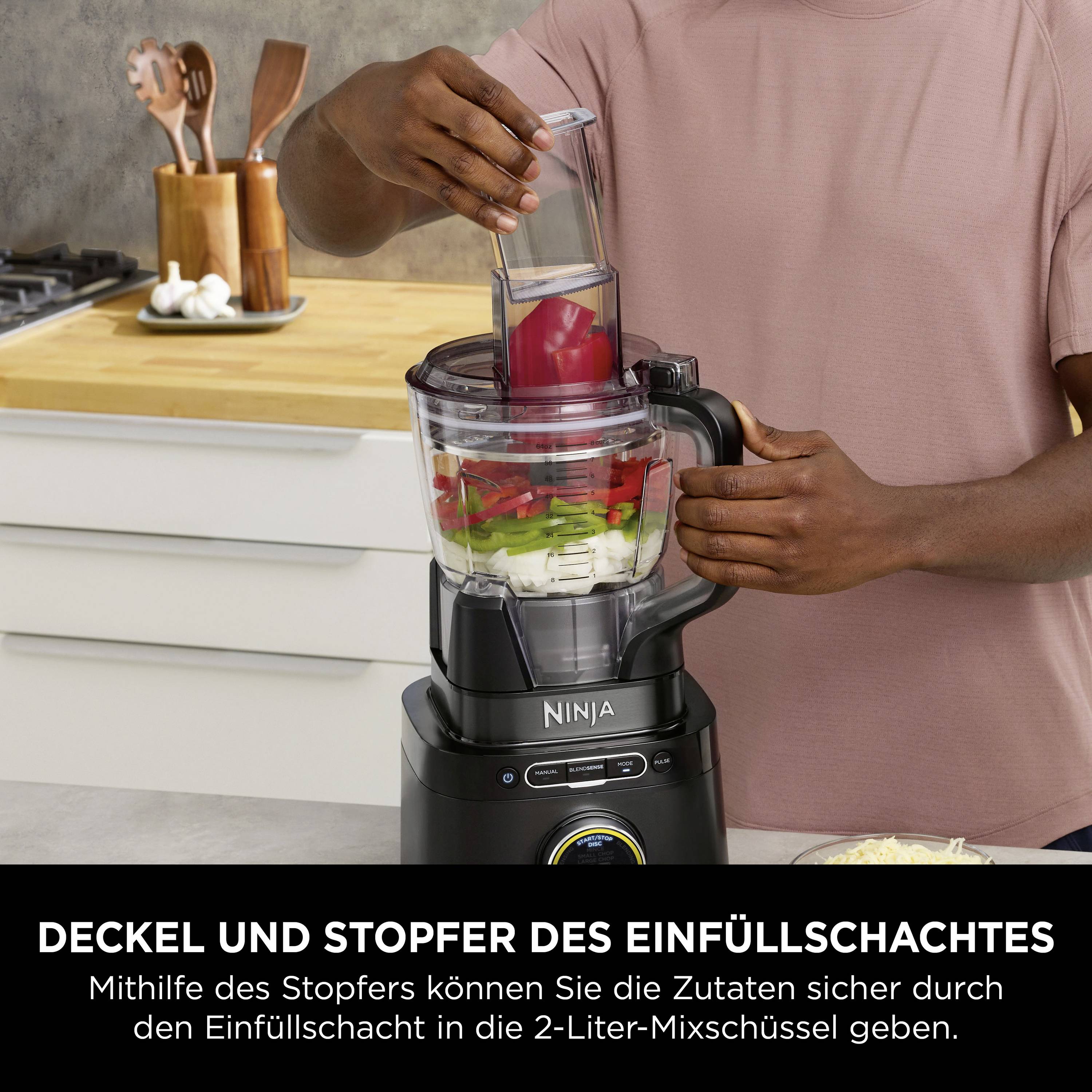 Ein Mann steckt Zutaten in einen Mixer mit der Aufschrift 'Deckel und Stopfer des Einfüllschachtes'. Küche im Hintergrund.