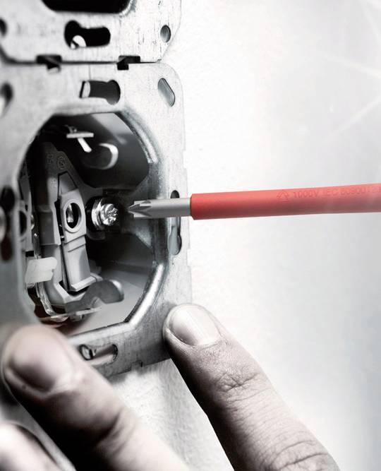 Eine Person schraubt mit einem roten Schraubendreher an einer Metallhalterung, möglicherweise eine Steckdose oder ein Schalter installierend.