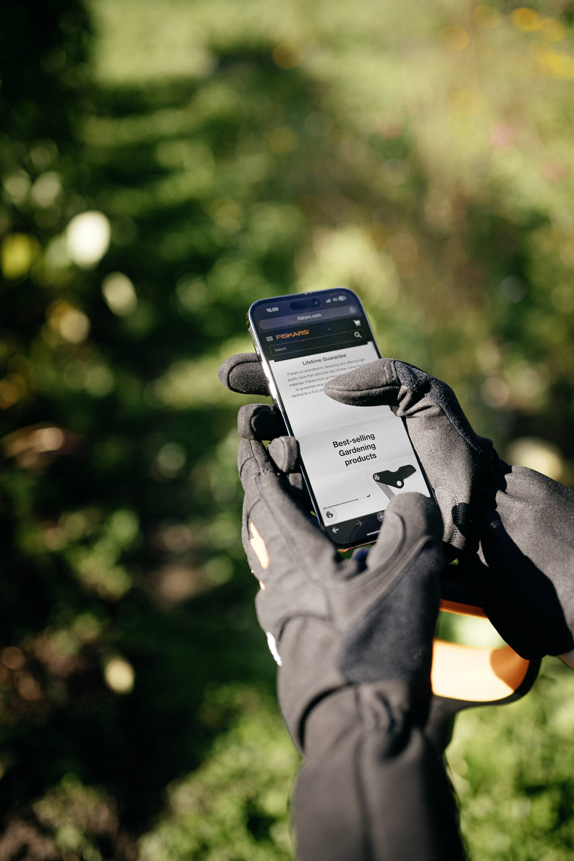 Hand in einem Garten hält ein Smartphone mit Garteninformationen auf dem Bildschirm. Person trägt schwarze Handschuhe.