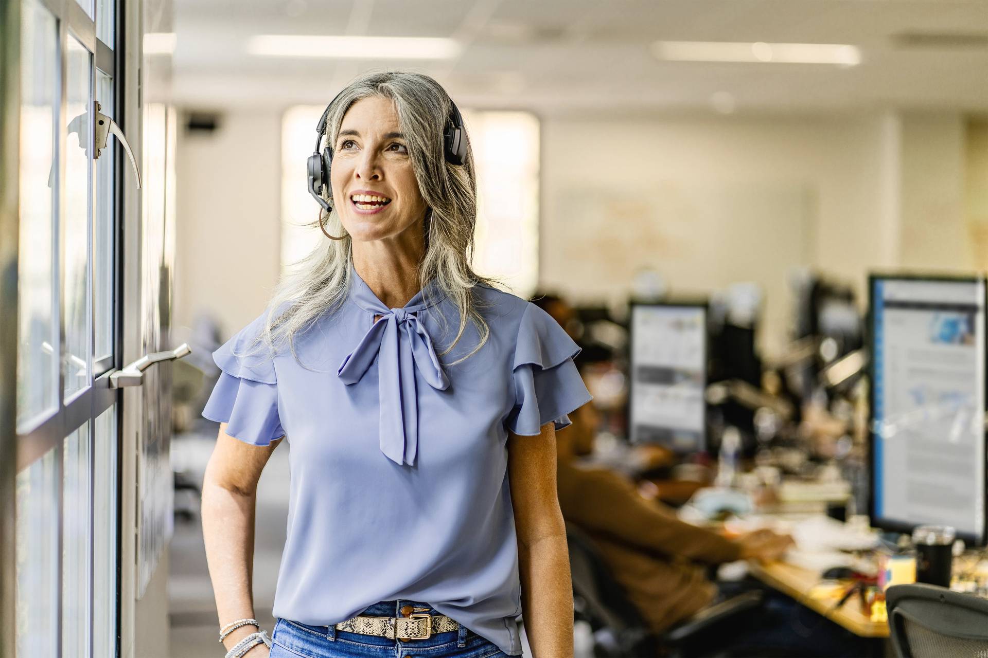 Eine Frau mit Kopfhörern lächelt und schaut aus einem Bürofenster. Im Hintergrund sind verschwommen Arbeitsplätze zu sehen.