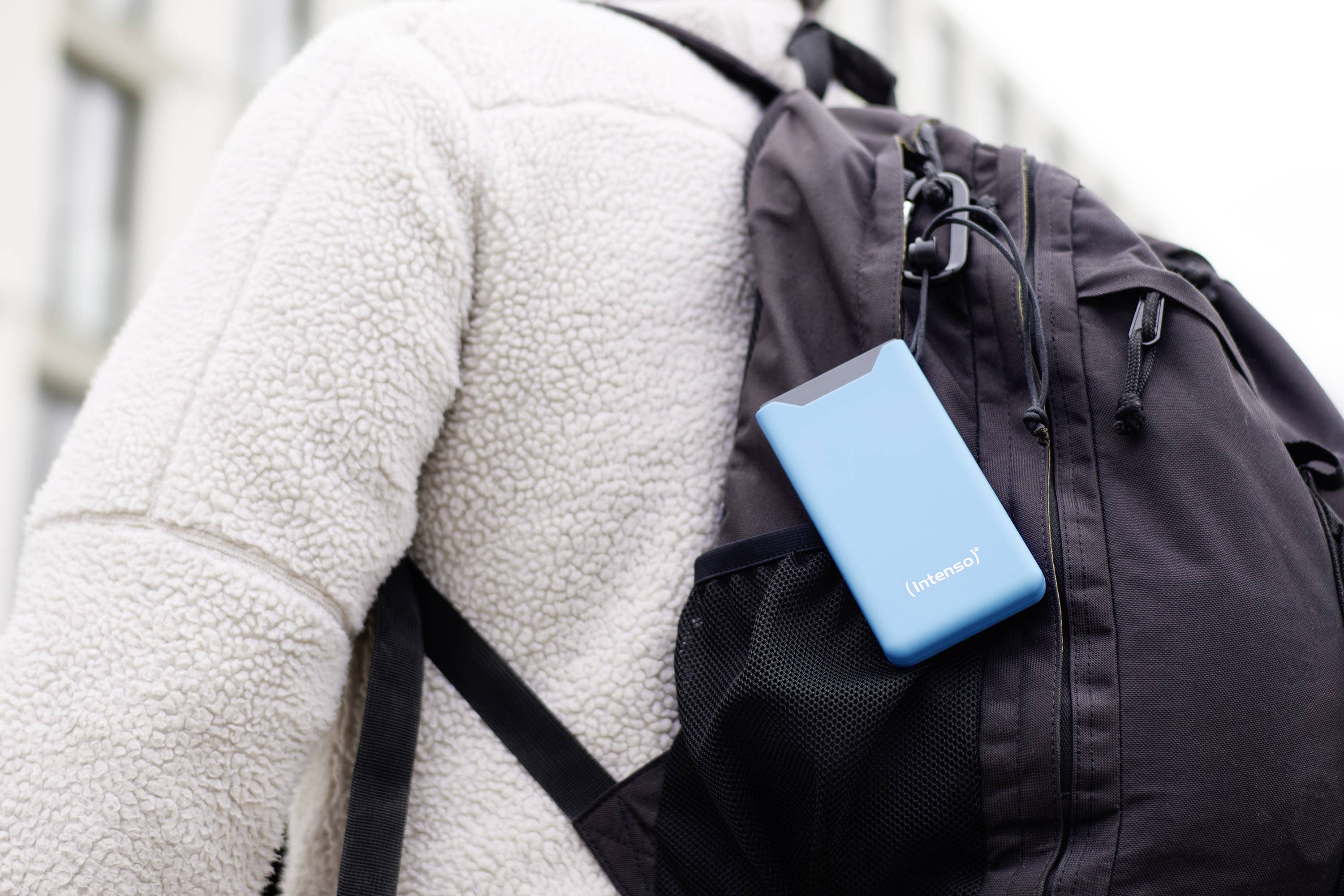 Eine Person mit weißer Jacke trägt einen schwarzen Rucksack, an dem ein blaues tragbares Gerät in einer Außentasche befestigt ist.